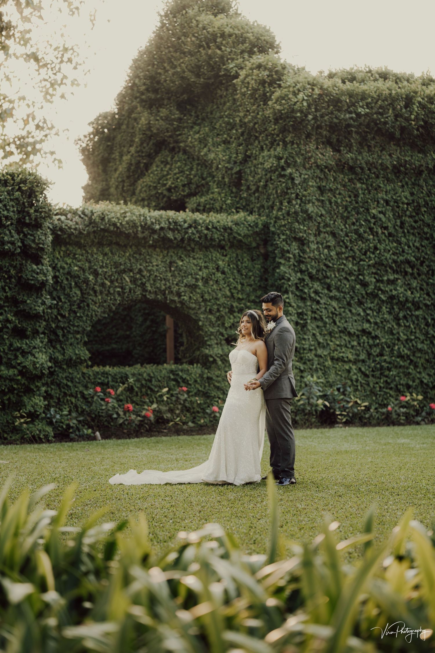 A couple embraces on a lush lawn surrounded by towering ivy-covered garden walls and flowering plants on their wedding day.
