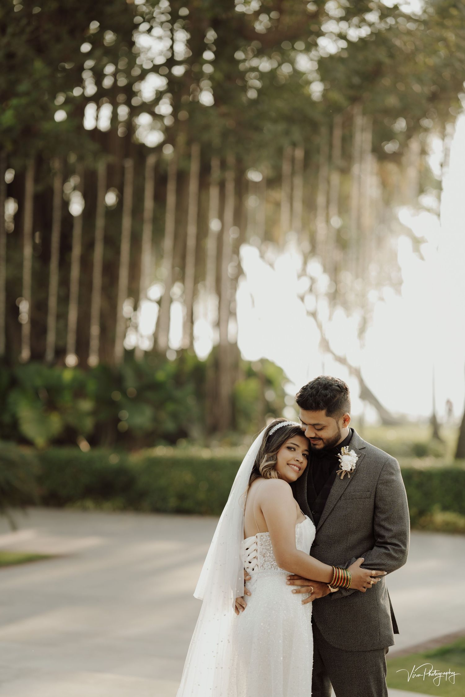 A romantic wedding couple embraces on a tree-lined pathway during golden hour sunset.