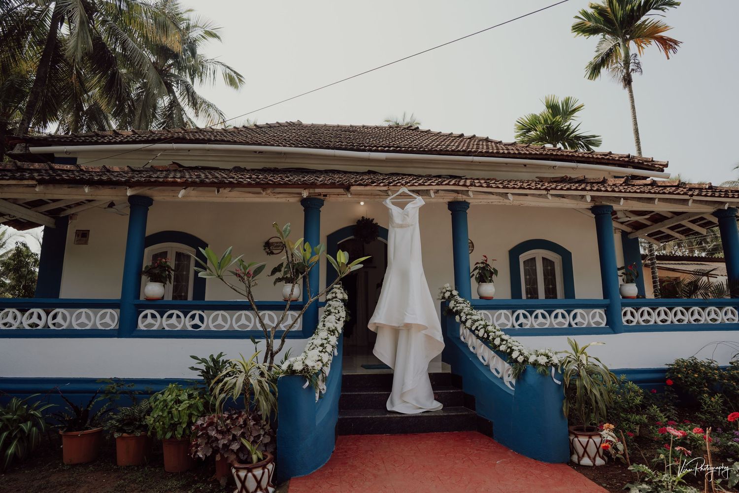 A white wedding dress hangs in the doorway of a Portuguese colonial style house with blue railings and palm trees in Goa.
