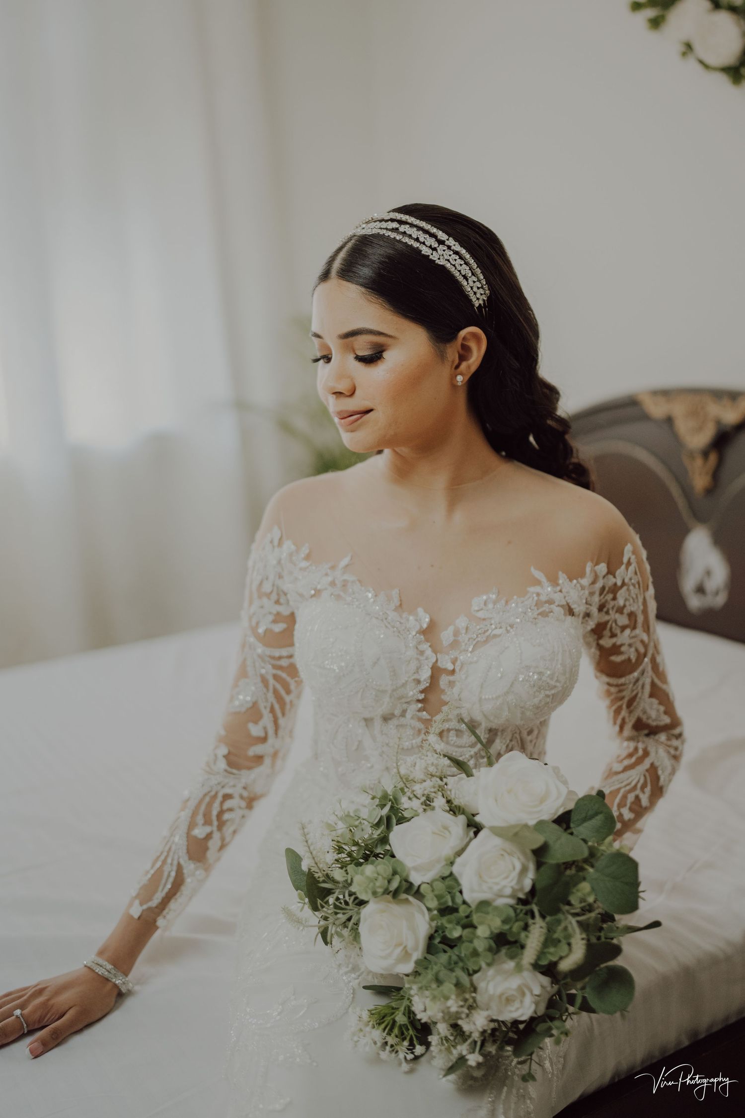 A bride in a lace long-sleeve wedding gown with an off-shoulder neckline holds a white and green bouquet while sitting.