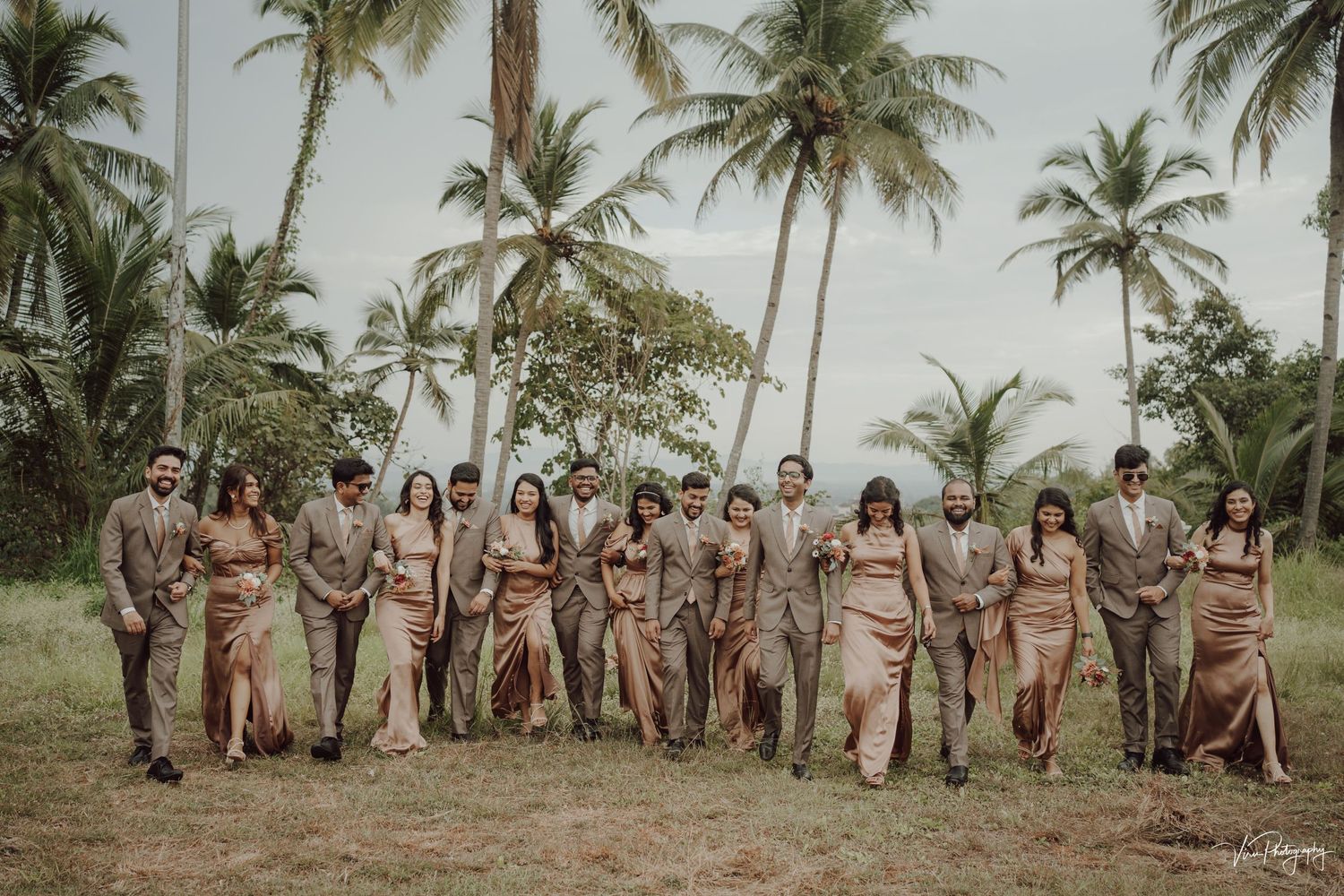 Wedding party standing together beneath tall palm trees on a tropical beach.