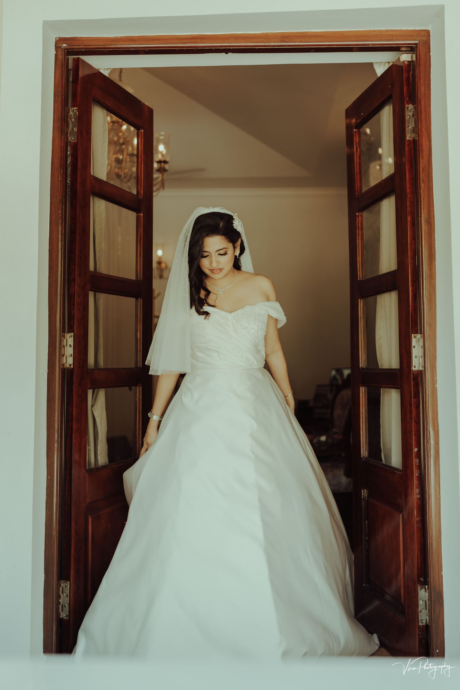 An elegant white off-shoulder wedding gown is showcased through open wooden double doors in a classic setting.