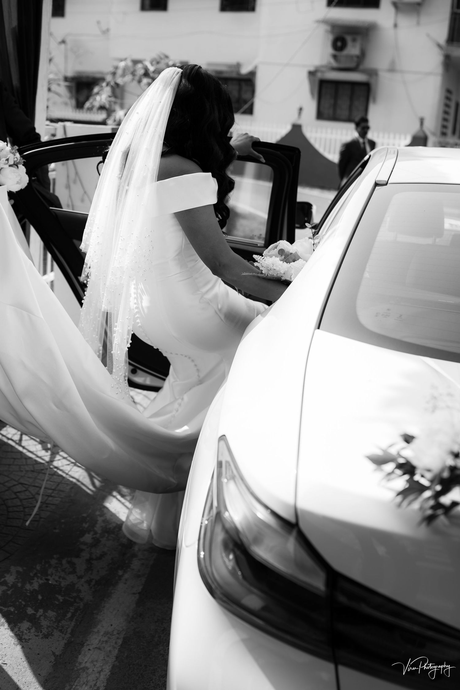 A bride in a flowing white dress enters a white luxury car in black and white photography.