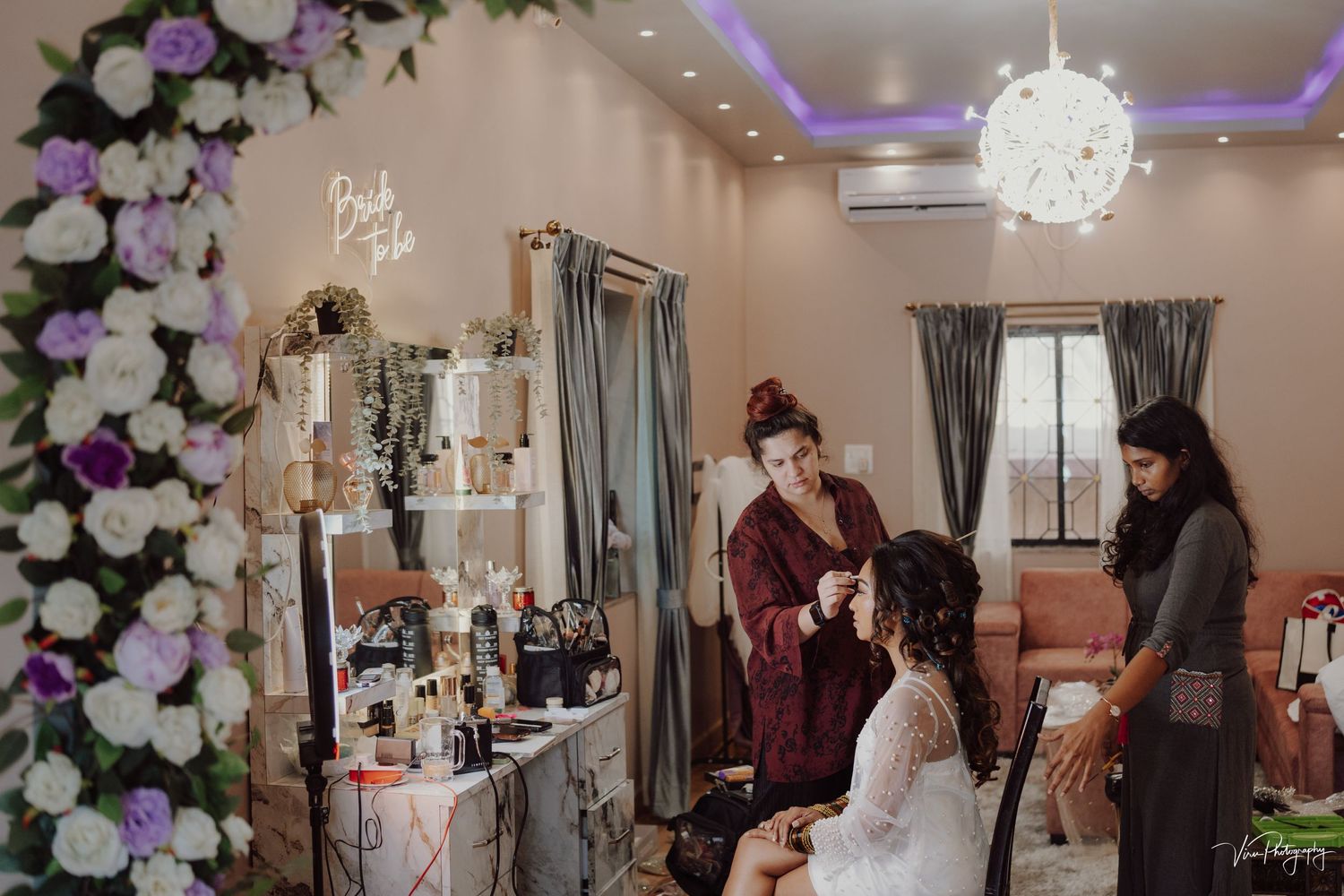 Wedding preparation scene in a room with string lights showing makeup tables, floral decorations and soft lighting details.