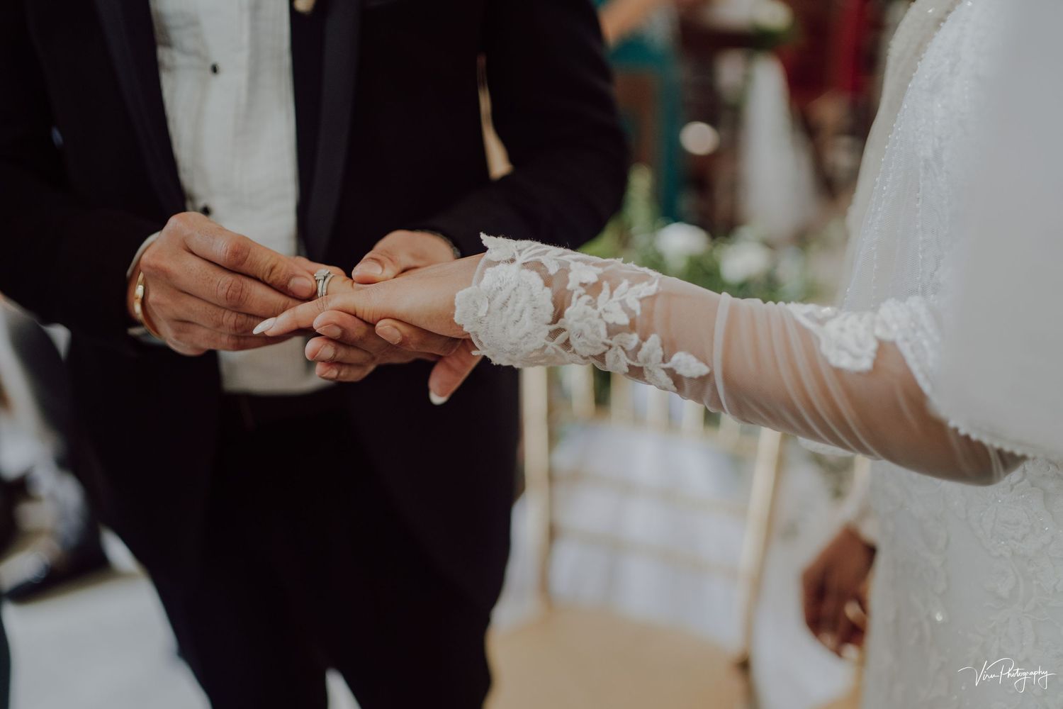 Close-up of hands exchanging wedding rings during an indoor ceremony with delicate lace sleeve details.