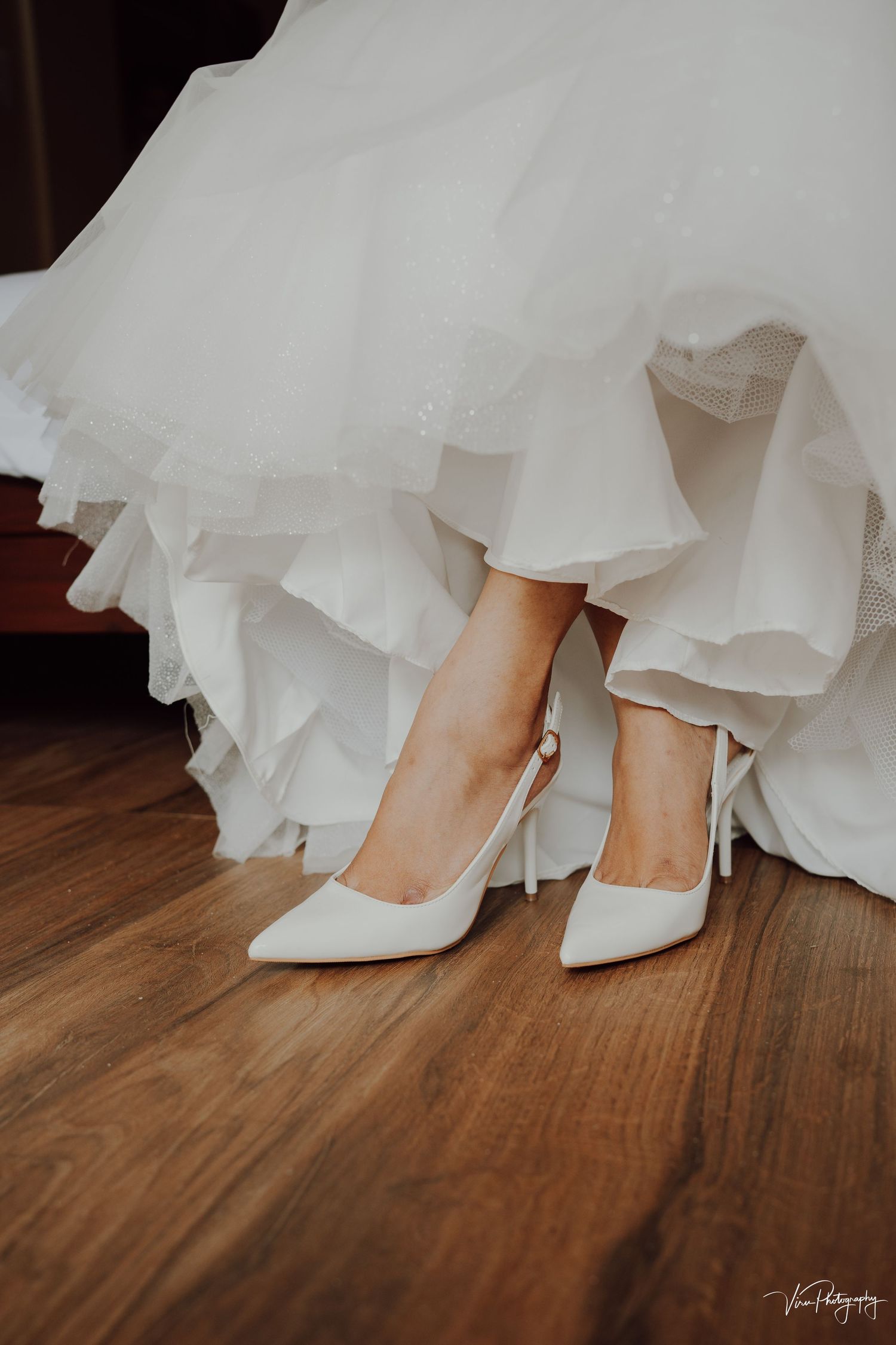Elegant white bridal shoes peek out beneath layers of flowing tulle wedding dress on hardwood floor.