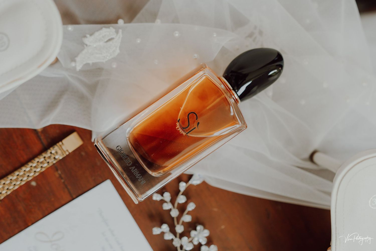 A glass perfume bottle laying on a white fabric surface next to a brown leather item and delicate white flowers.