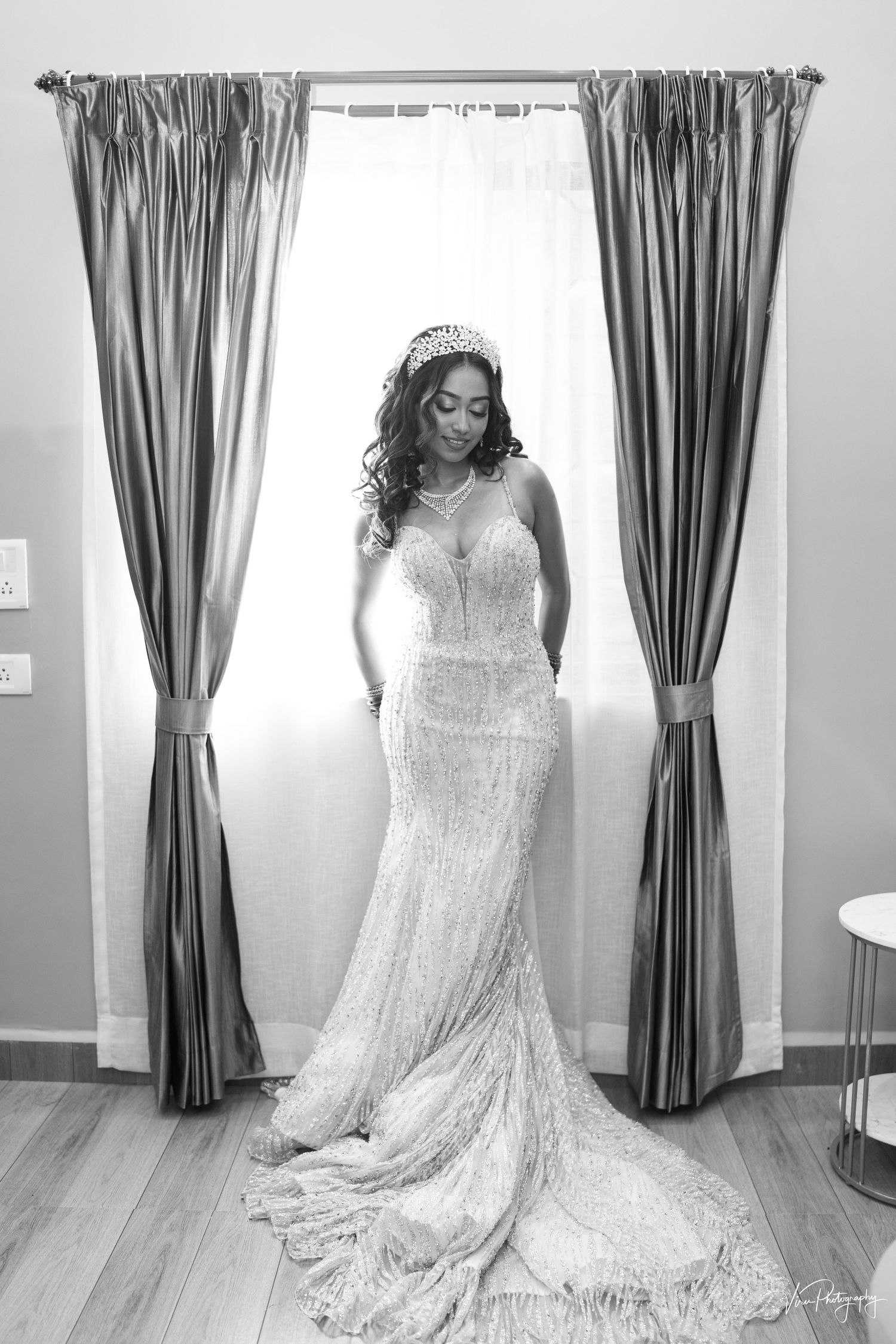 A bride wearing a sparkling white mermaid style wedding gown stands elegantly between grey curtains in a natural-lit room.