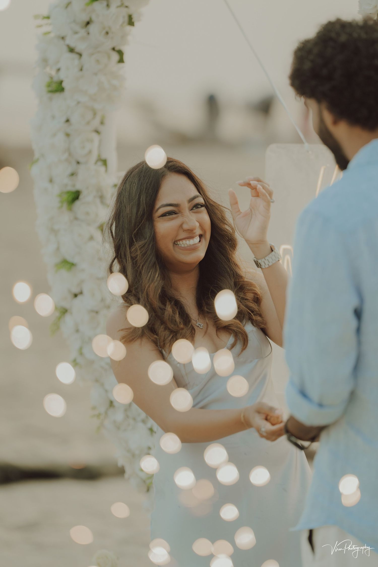 A couple shares a joyful moment under white floral decorations and twinkling bokeh lights at an outdoor celebration.