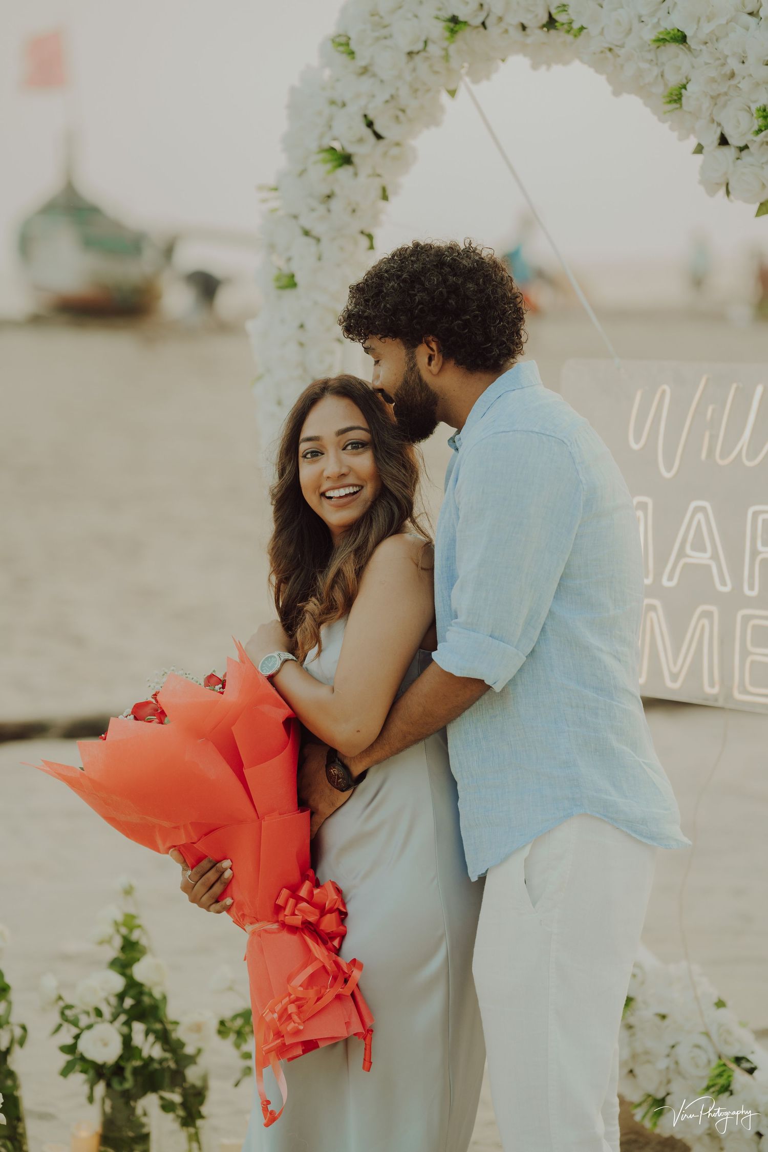A romantic beach proposal scene with white floral arch and coral bouquet against a dreamy beige background.