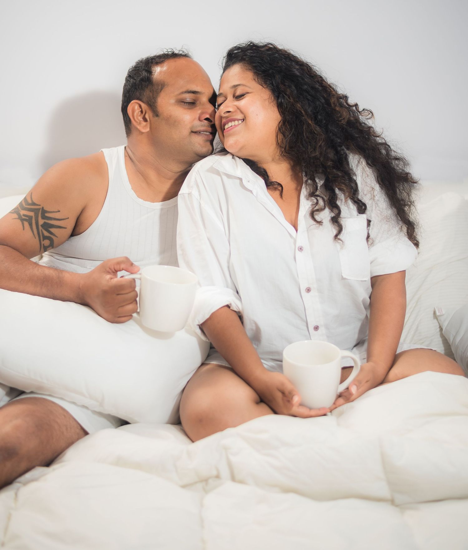 Couple sharing intimate morning moment in bed while drinking coffee and tea against white wall.