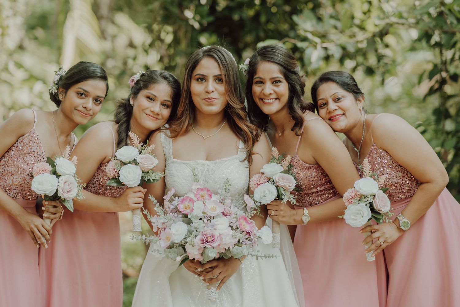 Wedding party wearing matching pink sequined dresses poses together holding white and pink floral bouquets in outdoor garden.