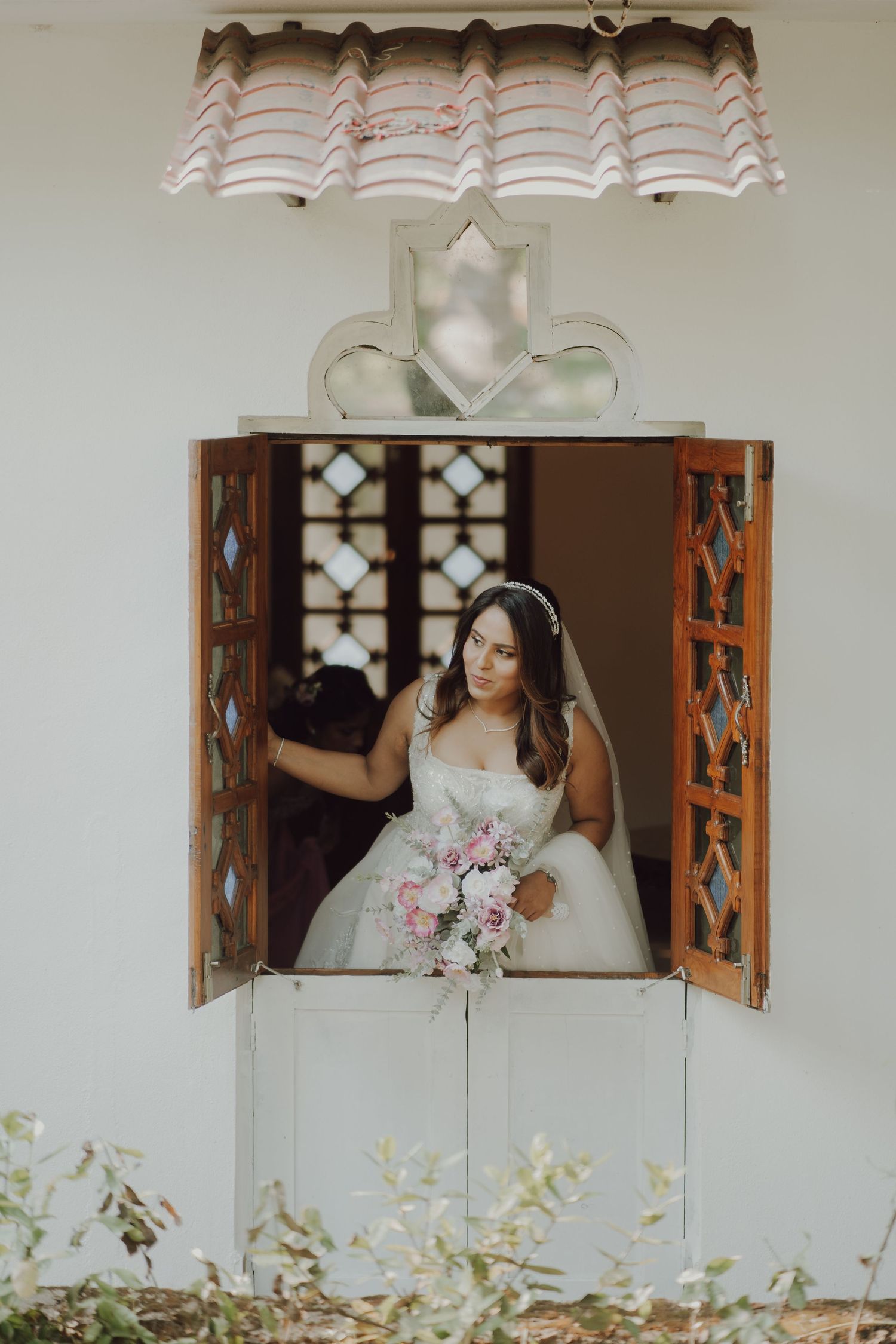 A bride in a white dress holds a pink bouquet while standing at a decorative window with ornate wooden shutters.