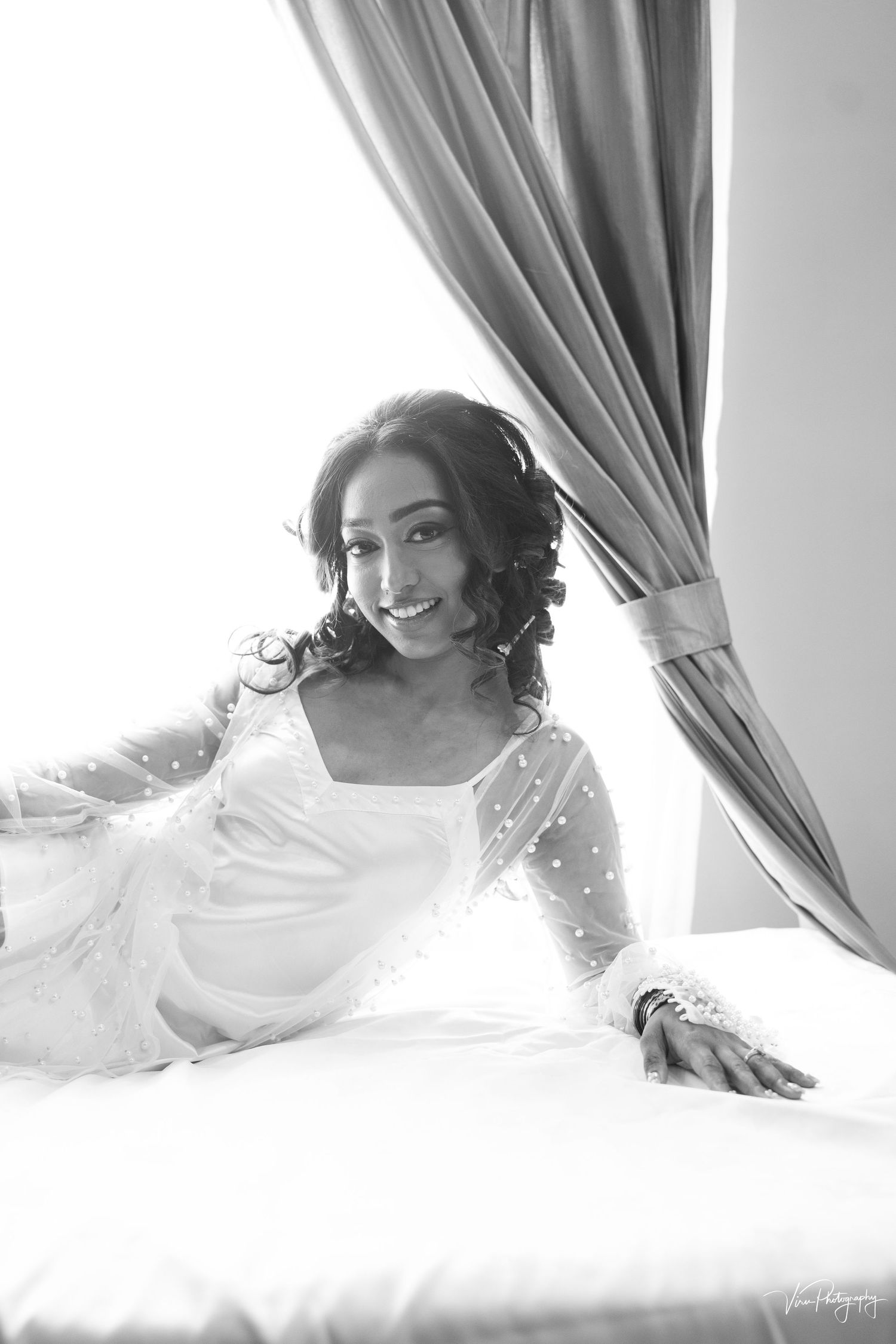 A black and white portrait of a bride in a white dress with sheer sleeves reclining by window drapes.