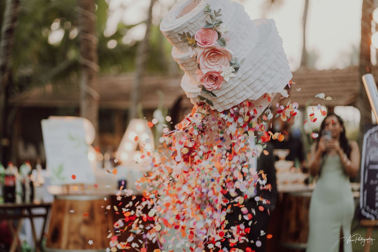 Bride and groom celebrate their outdoor wedding ceremony as pink and red confetti flutters through the sunlit air.