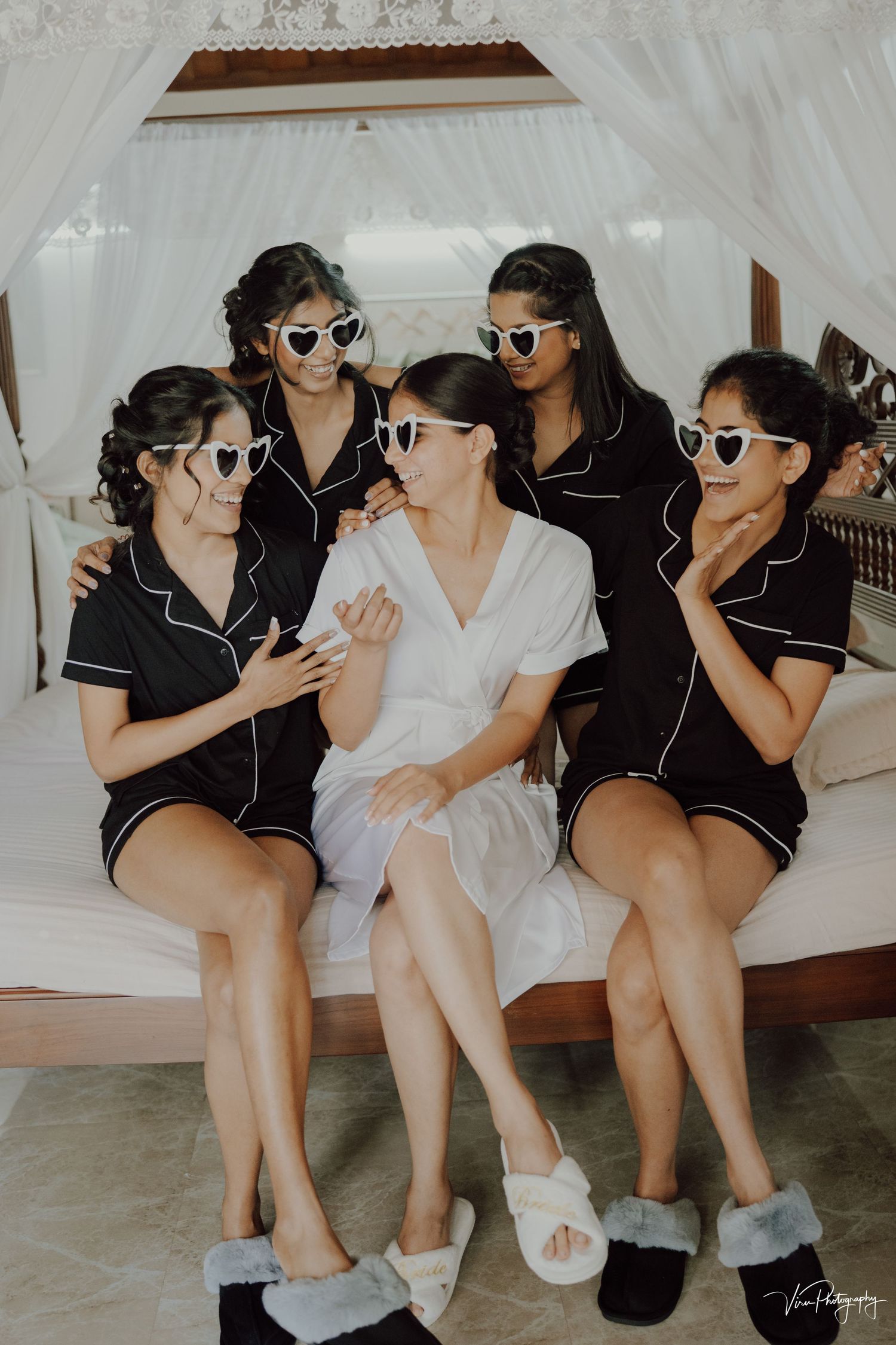 Wedding party wearing matching silk pajamas and sunglasses poses together on white canopy bed for fun photo shoot.