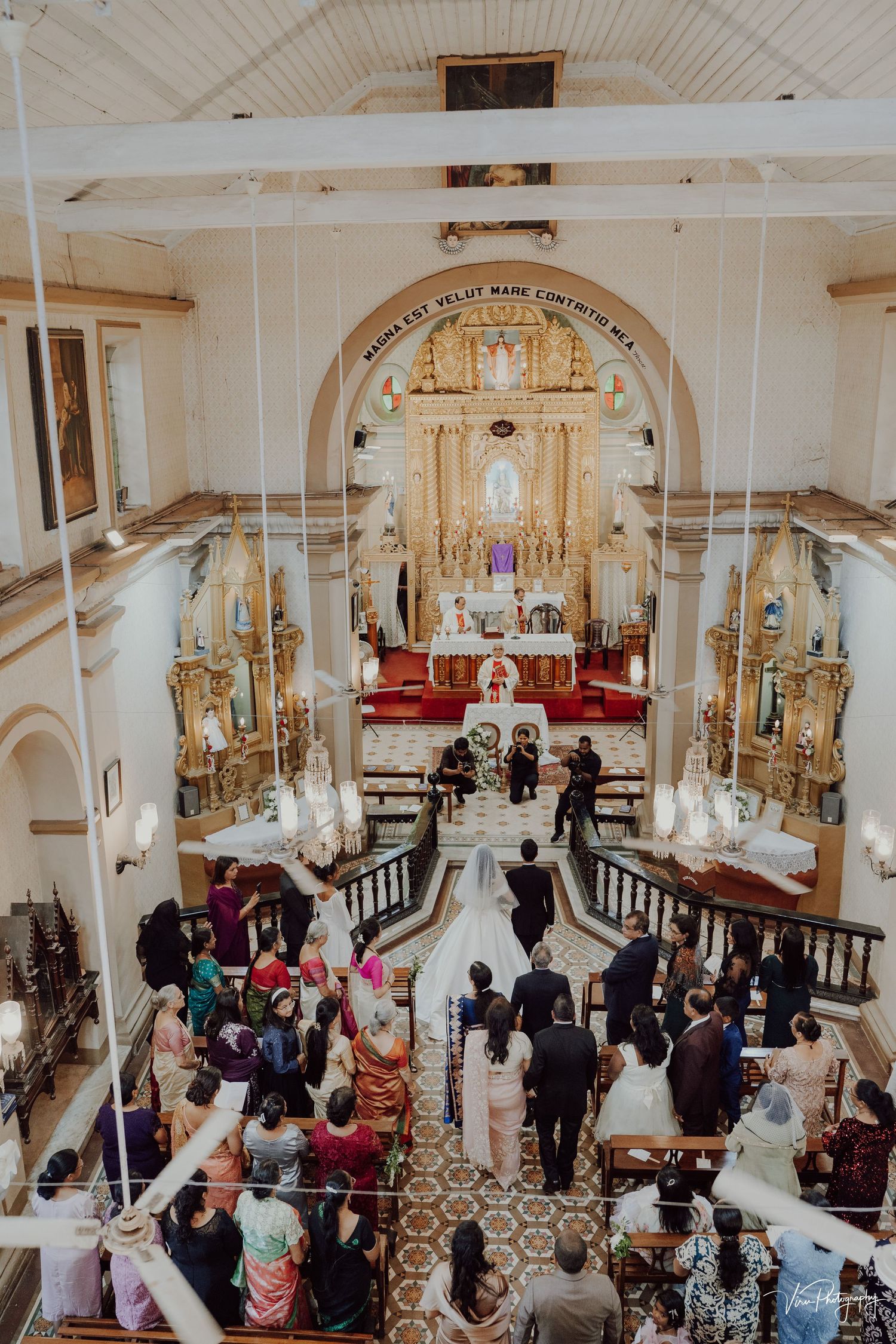 Aerial view of a wedding ceremony inside an ornate Catholic church with golden altar and guests gathered in the pews.