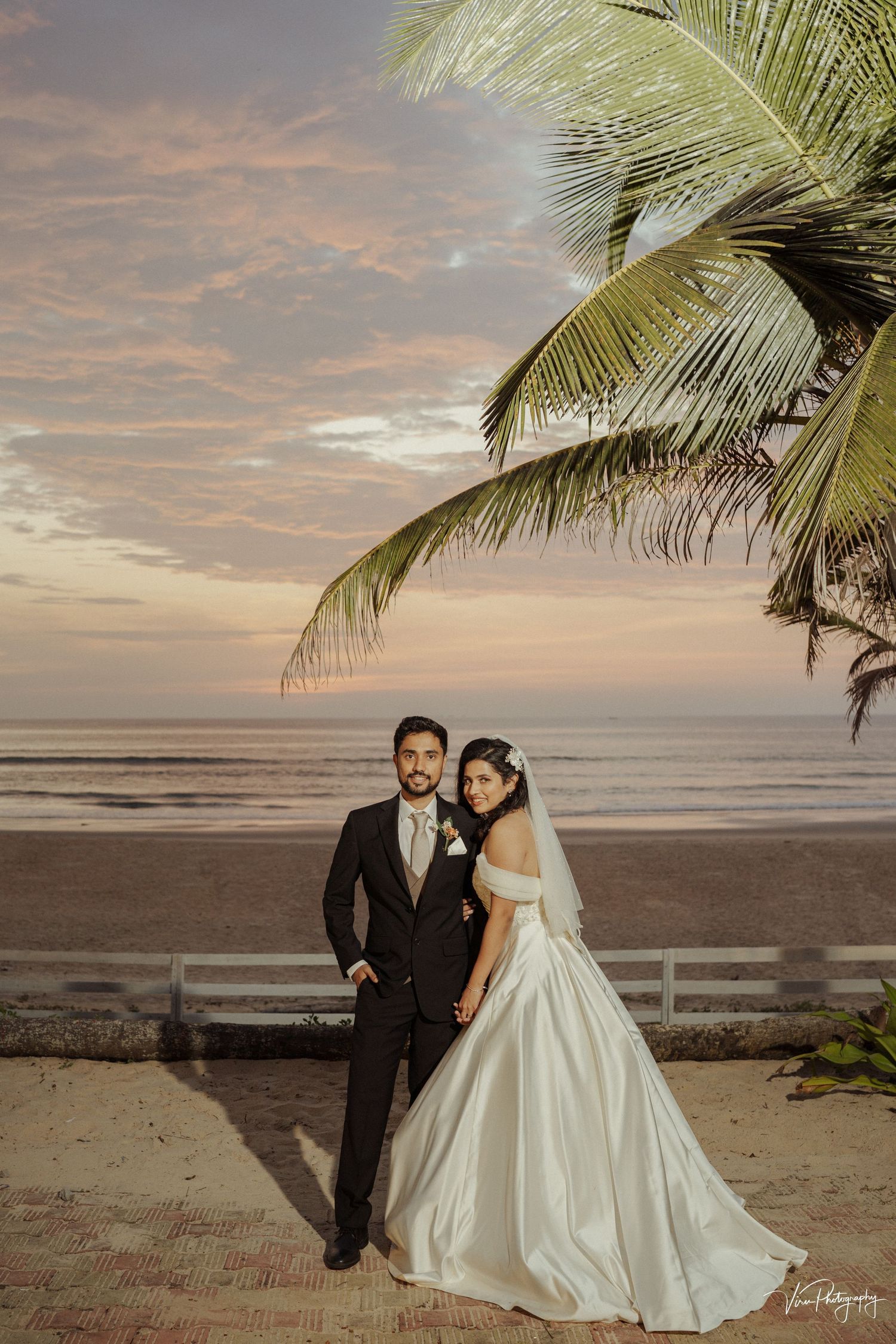 A couple poses for a sunset beach wedding photo with palm trees and ocean backdrop at golden hour.