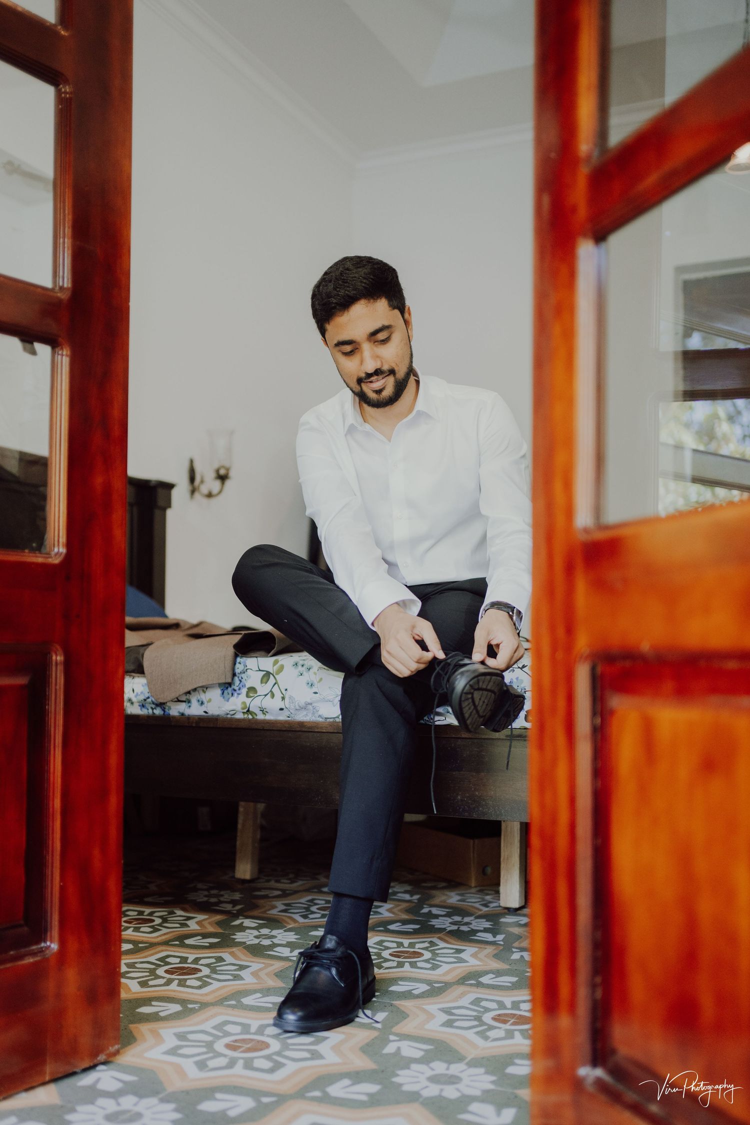 A well-dressed individual sits casually with crossed legs on a bench with ornate tiled flooring and rich wooden doorframes.