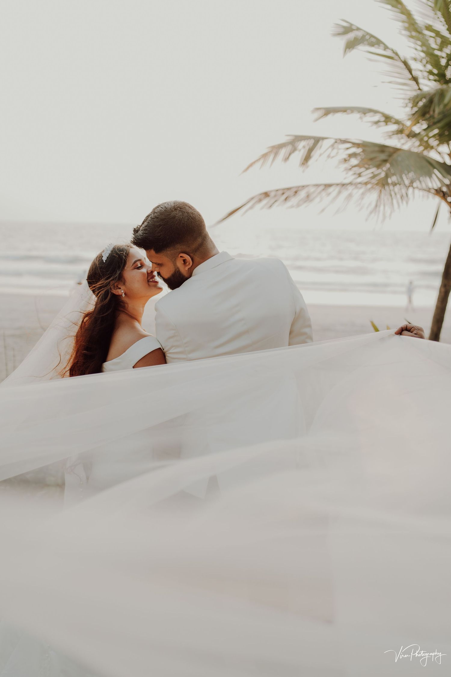 Romantic beach wedding couple embracing under a palm tree with flowing white veil catching the breeze at sunset.