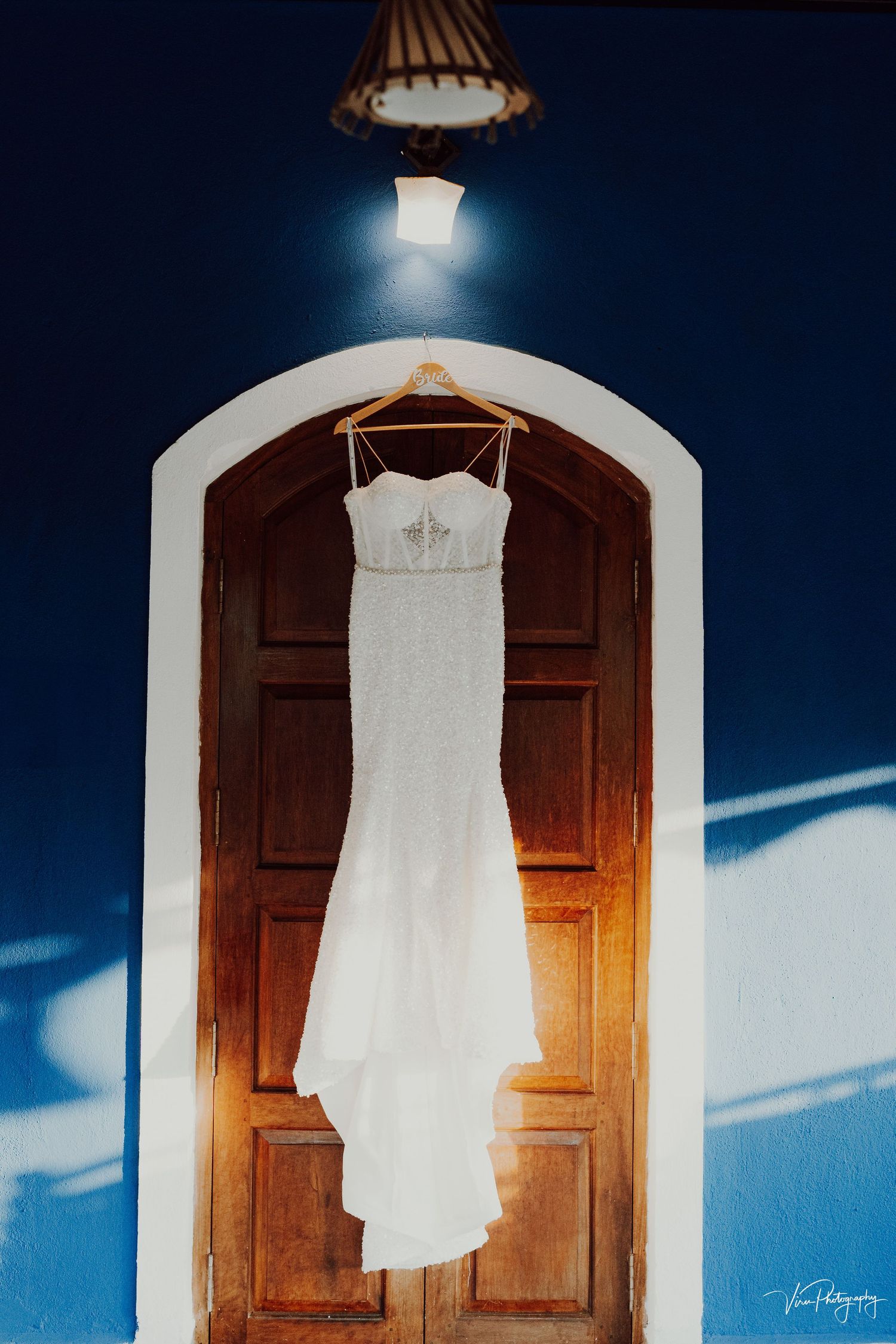 A white wedding dress hangs elegantly against dark wooden doors, illuminated by natural light and an overhead lamp.