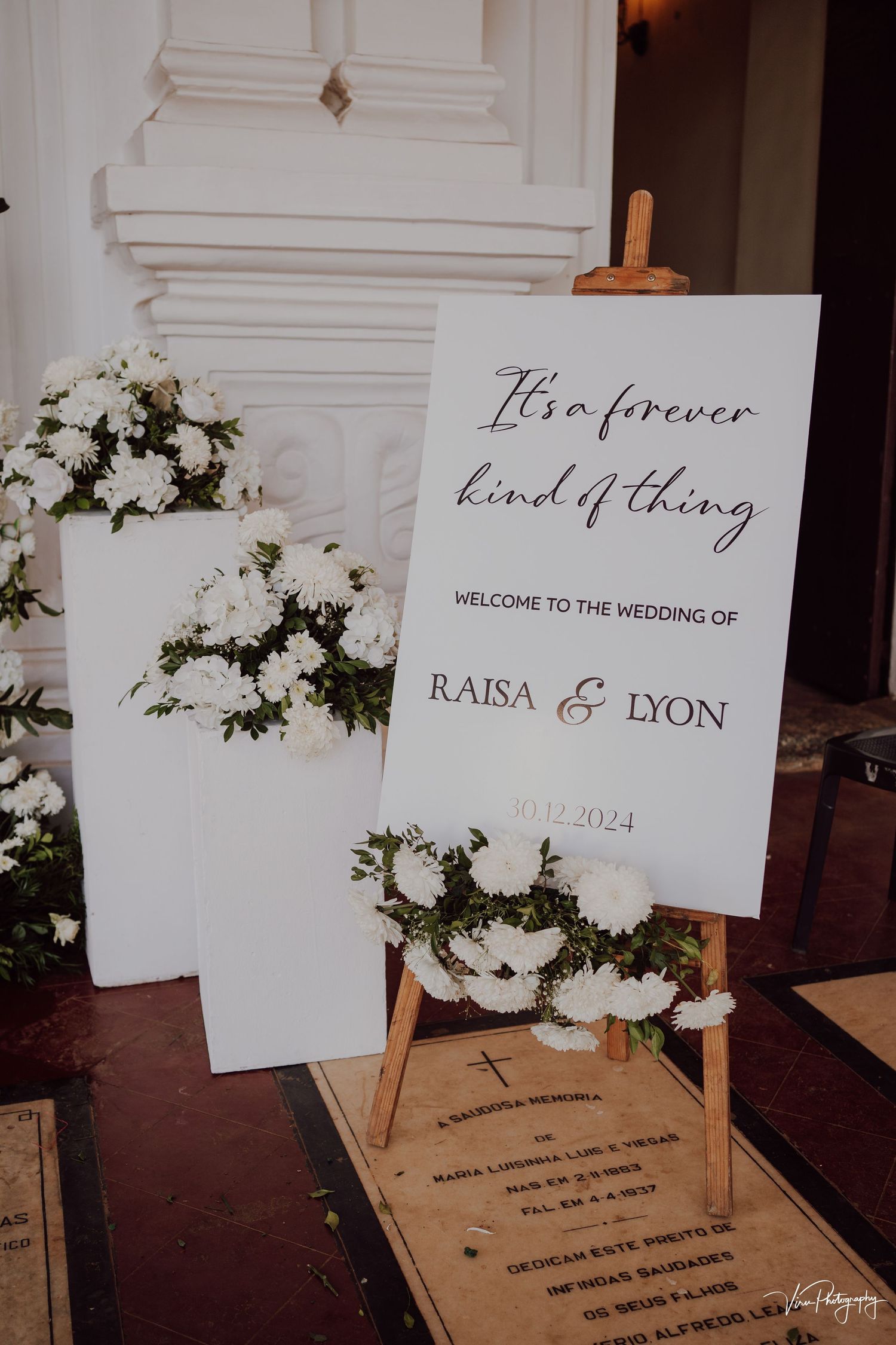Elegant wedding welcome sign with white floral arrangements and wooden easel displaying 'It's a forever kind of thing' message.