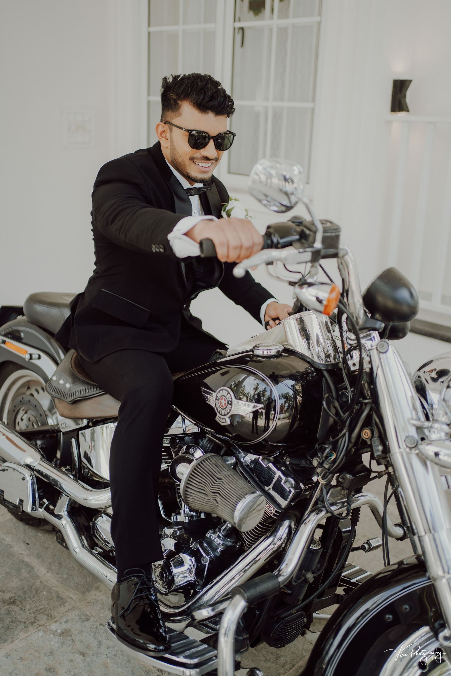A stylishly dressed person in black suit and sunglasses sits on a sleek black and chrome motorcycle.