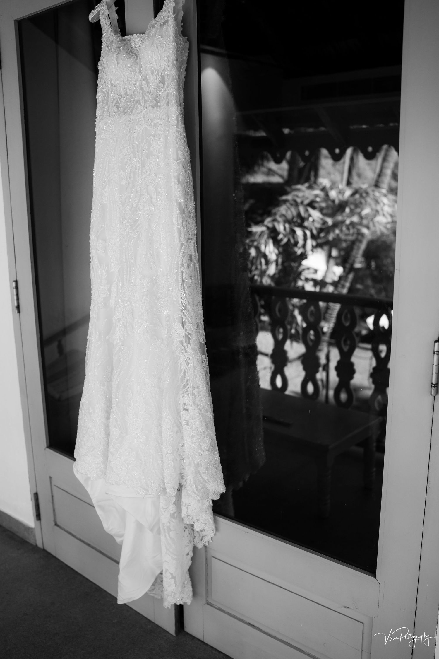 A delicate white lace wedding dress hangs elegantly against a reflective glass door in black and white photography.