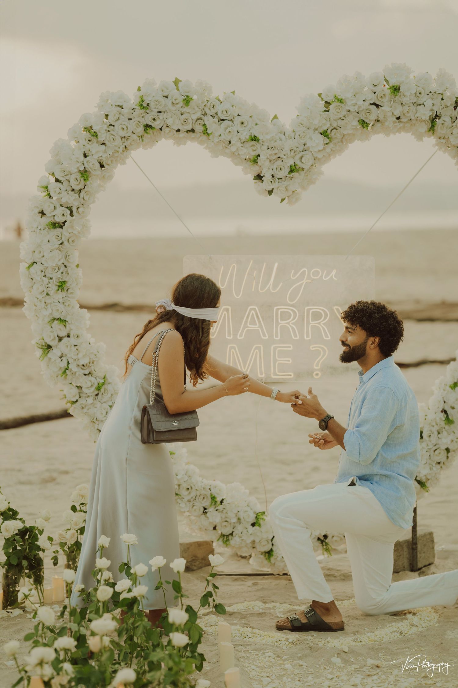 A romantic beach proposal setup with a white floral arch and 