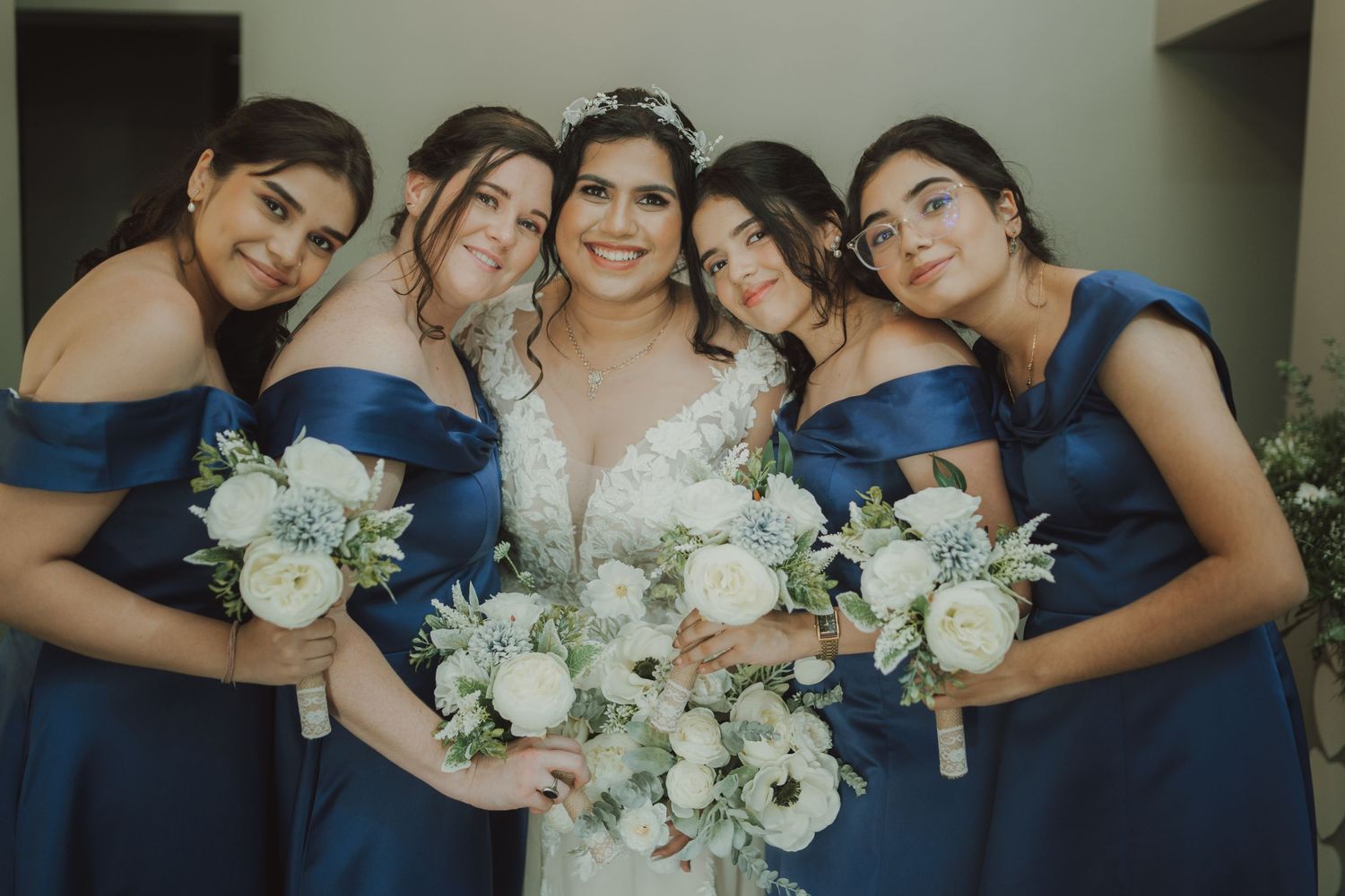 Bridal party in matching navy blue dresses hold white floral bouquets while posing together for wedding photo.