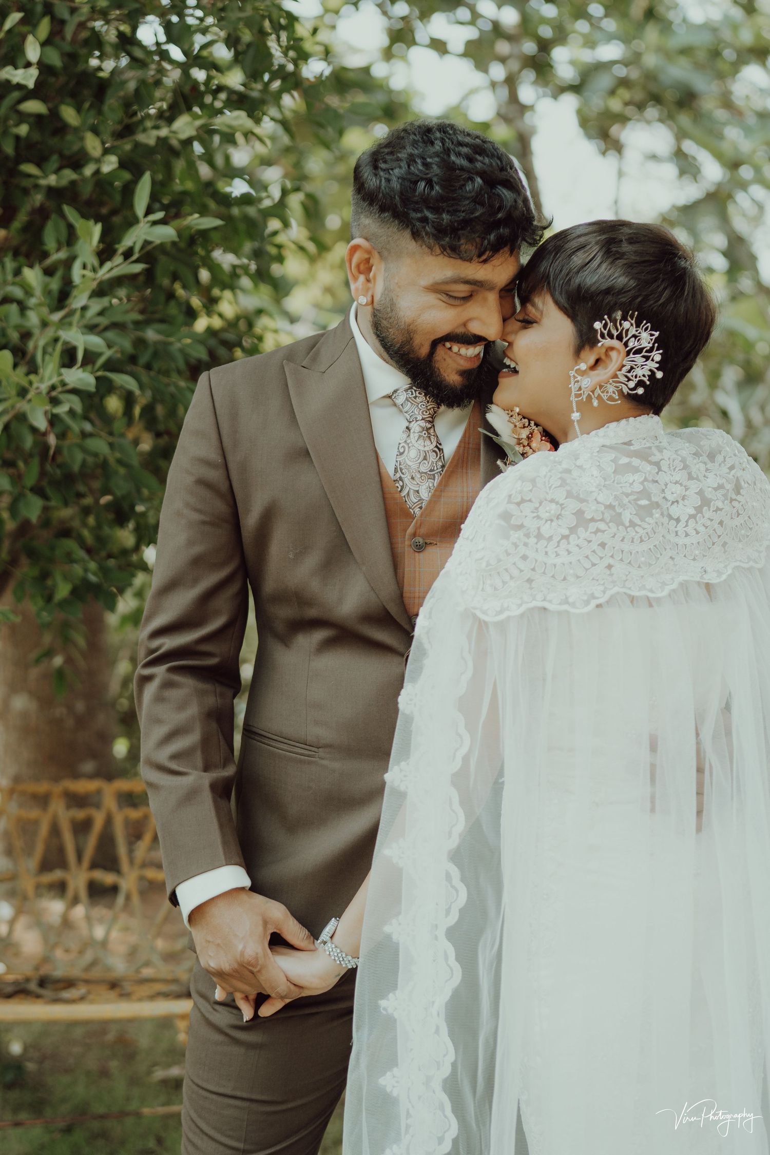 Newlywed couple shares an intimate moment outdoors, with bride in lace veil and groom in brown suit.