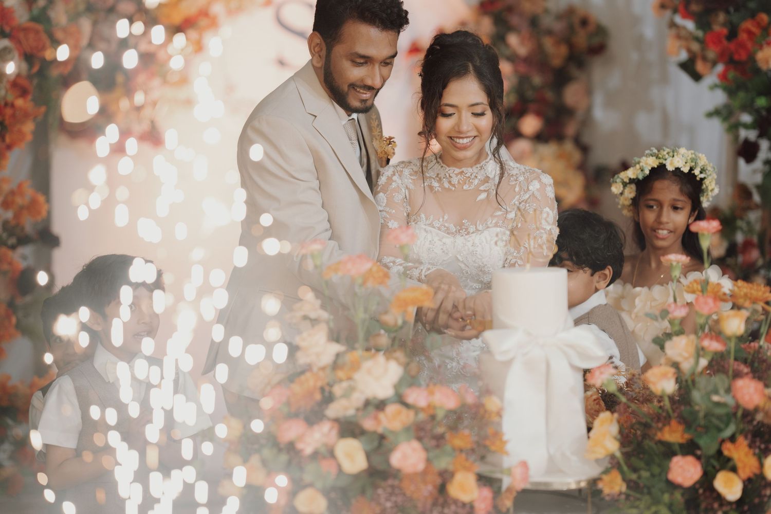 A bride and groom stand behind a white candle surrounded by orange and pink flowers, with sparkling lights and children beside them.