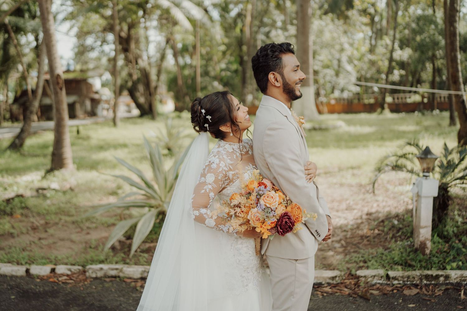 A bride in a white lace gown and veil embraces her groom in a beige suit, holding an orange bouquet in a lush tropical garden.