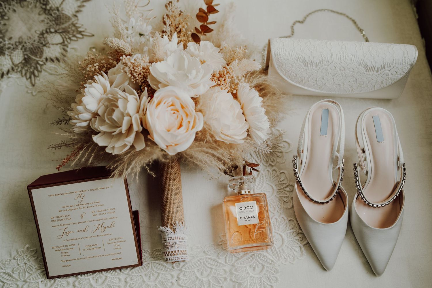 Wedding accessories including white heels, perfume, and a floral arrangement on a light surface.
