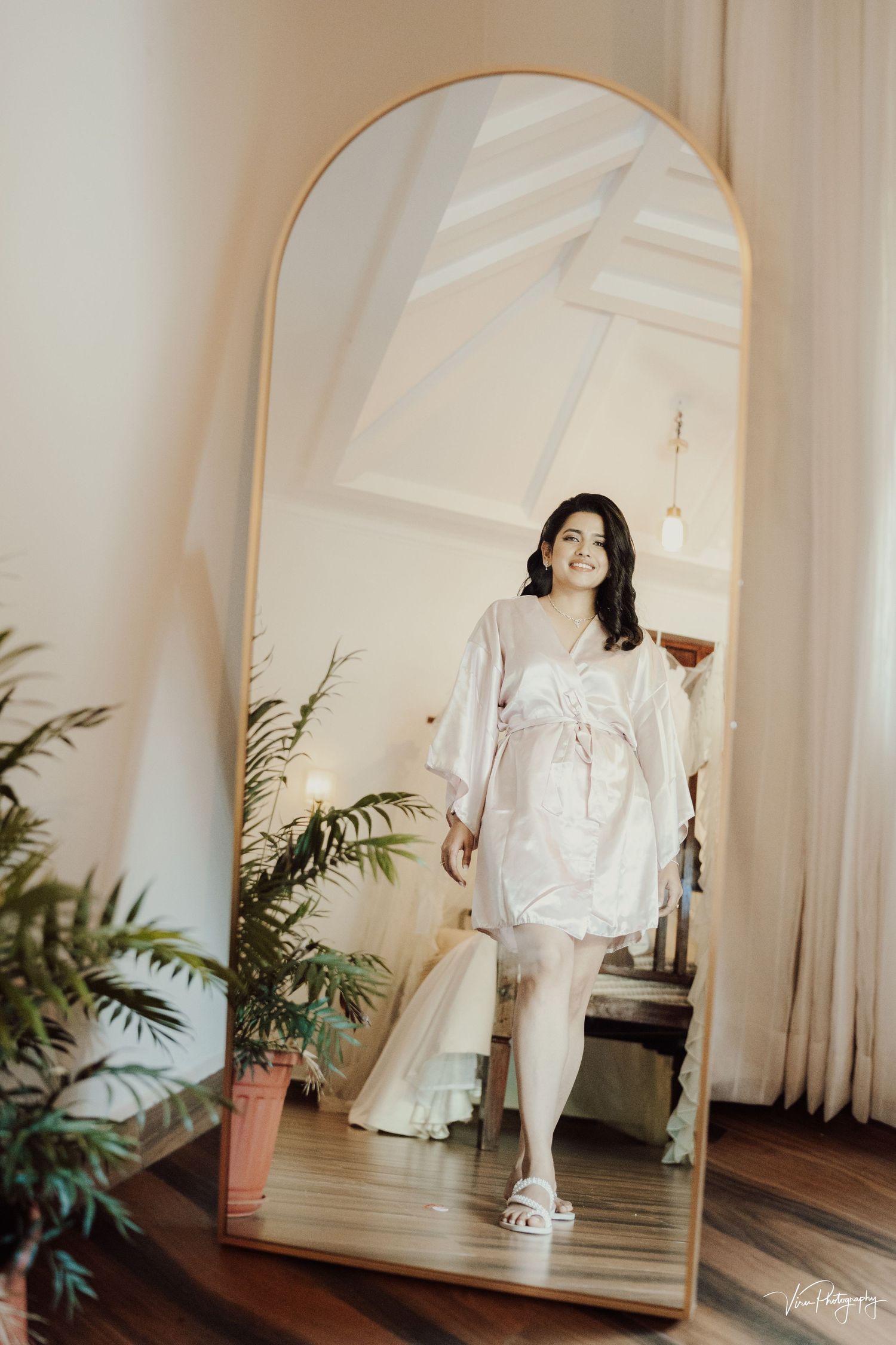 Mirror reflection shows a white silk robe and slippers against an elegant interior with palm plants and white curtains.