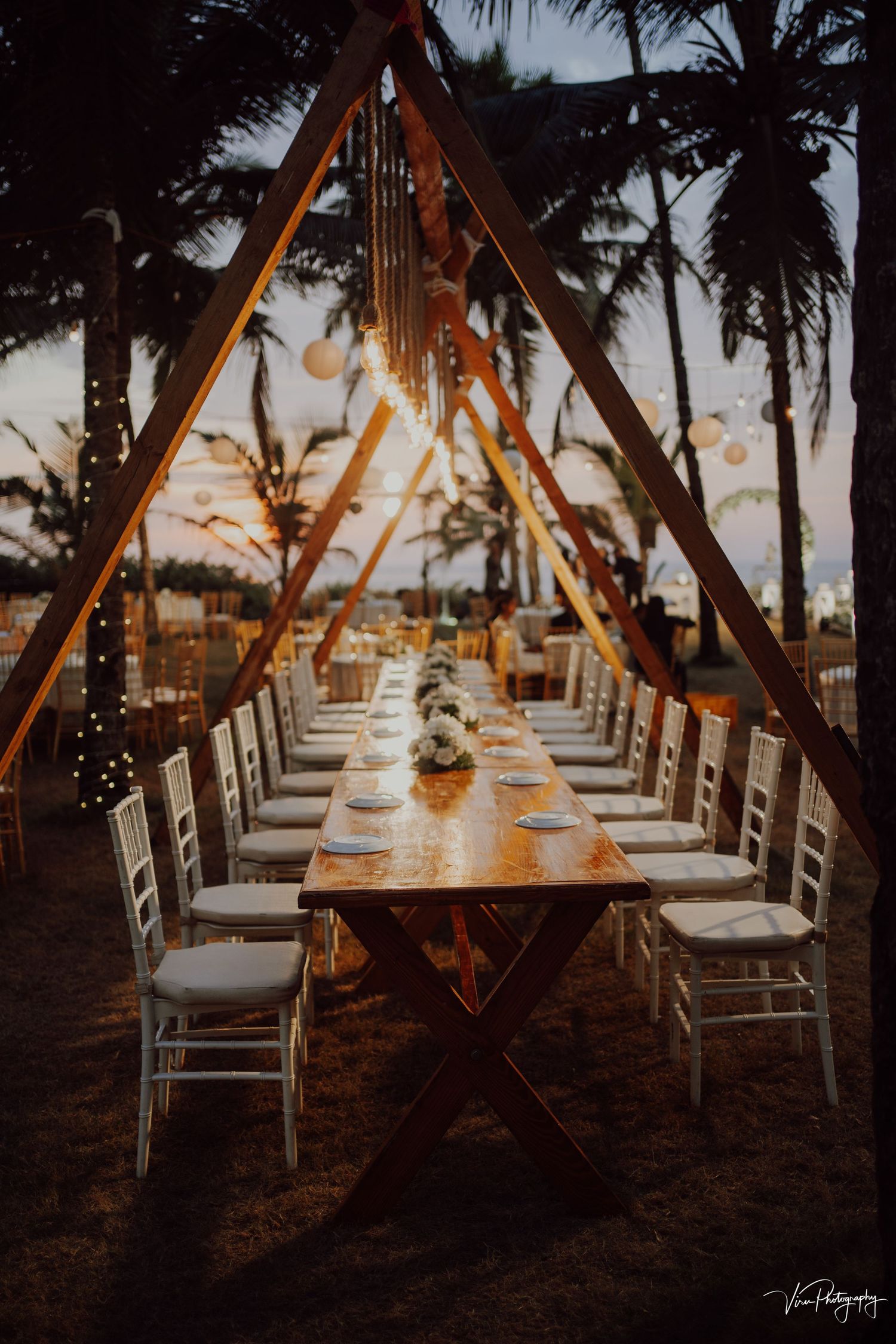 A rustic outdoor wedding reception table setup with string lights and palm trees silhouetted against a sunset sky.