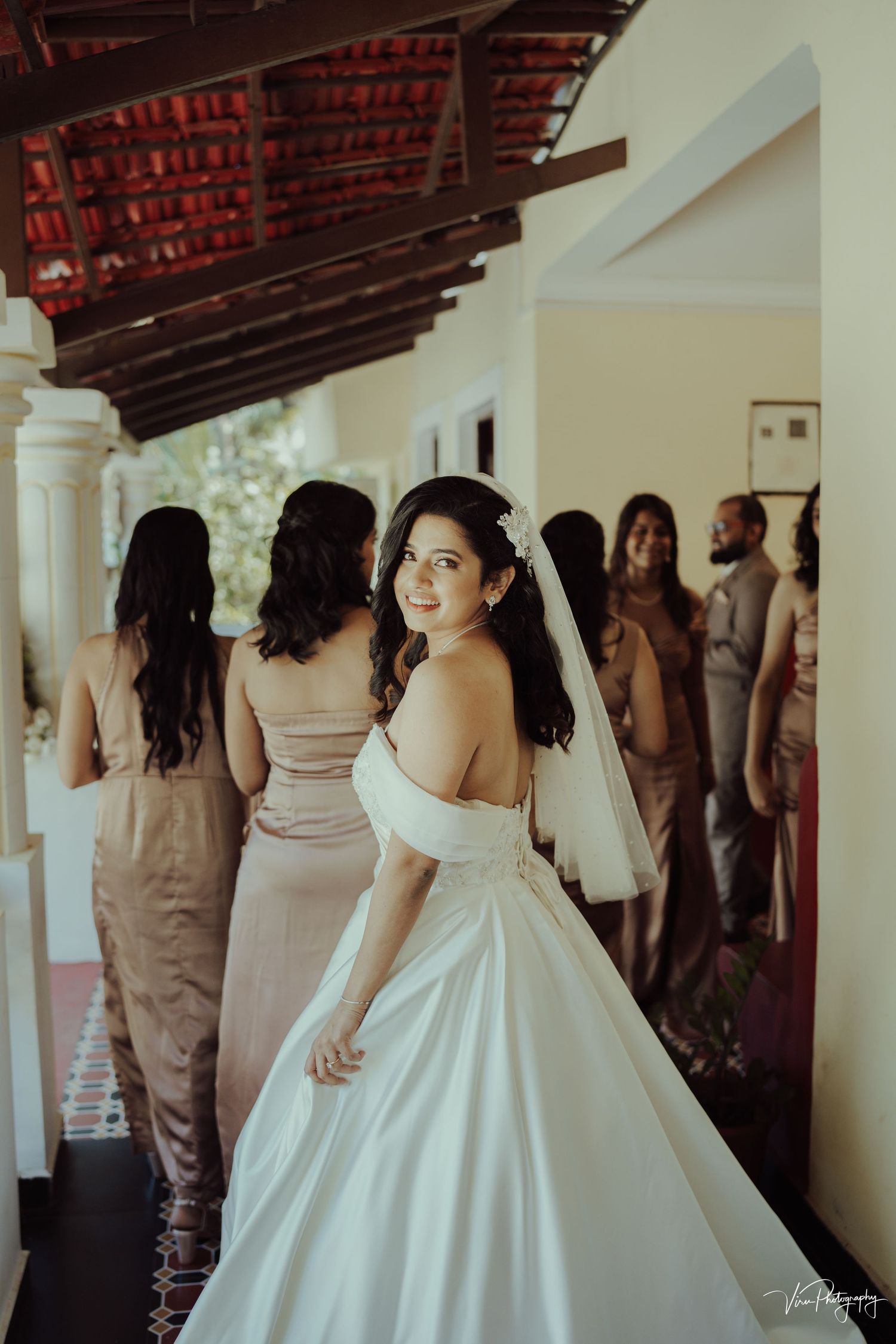 A bride in an elegant off-shoulder white ballgown stands on a covered porch with bridesmaids in blush dresses behind her.