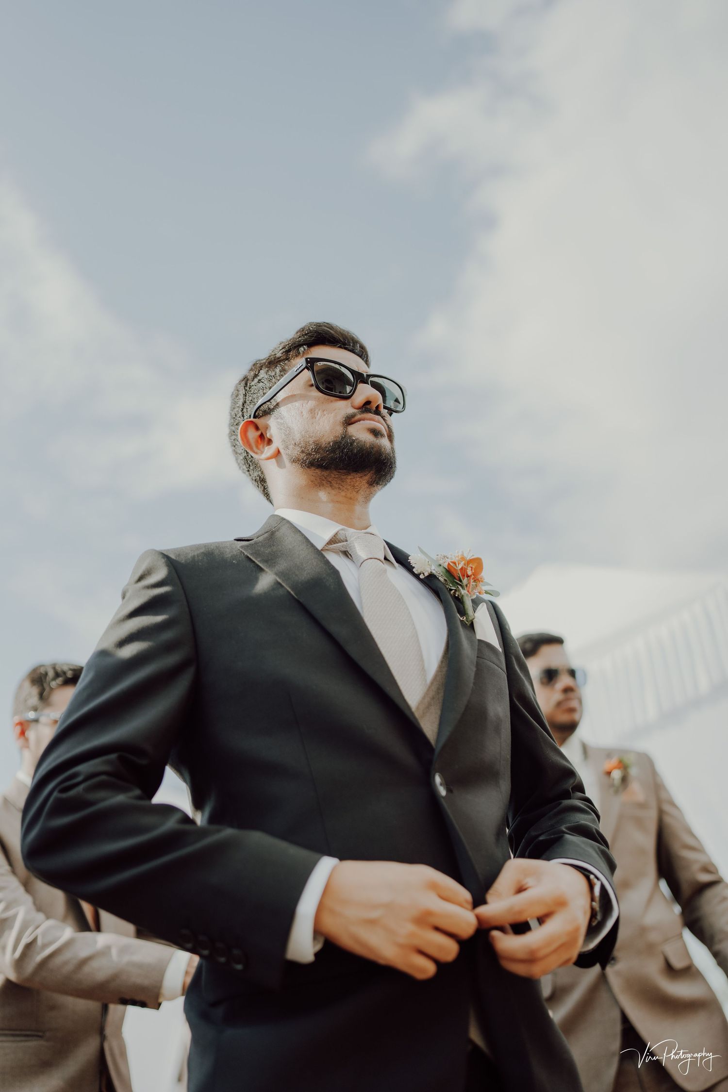 Stylish groomsman in dark suit and sunglasses poses confidently against cloudy sky during outdoor wedding ceremony.