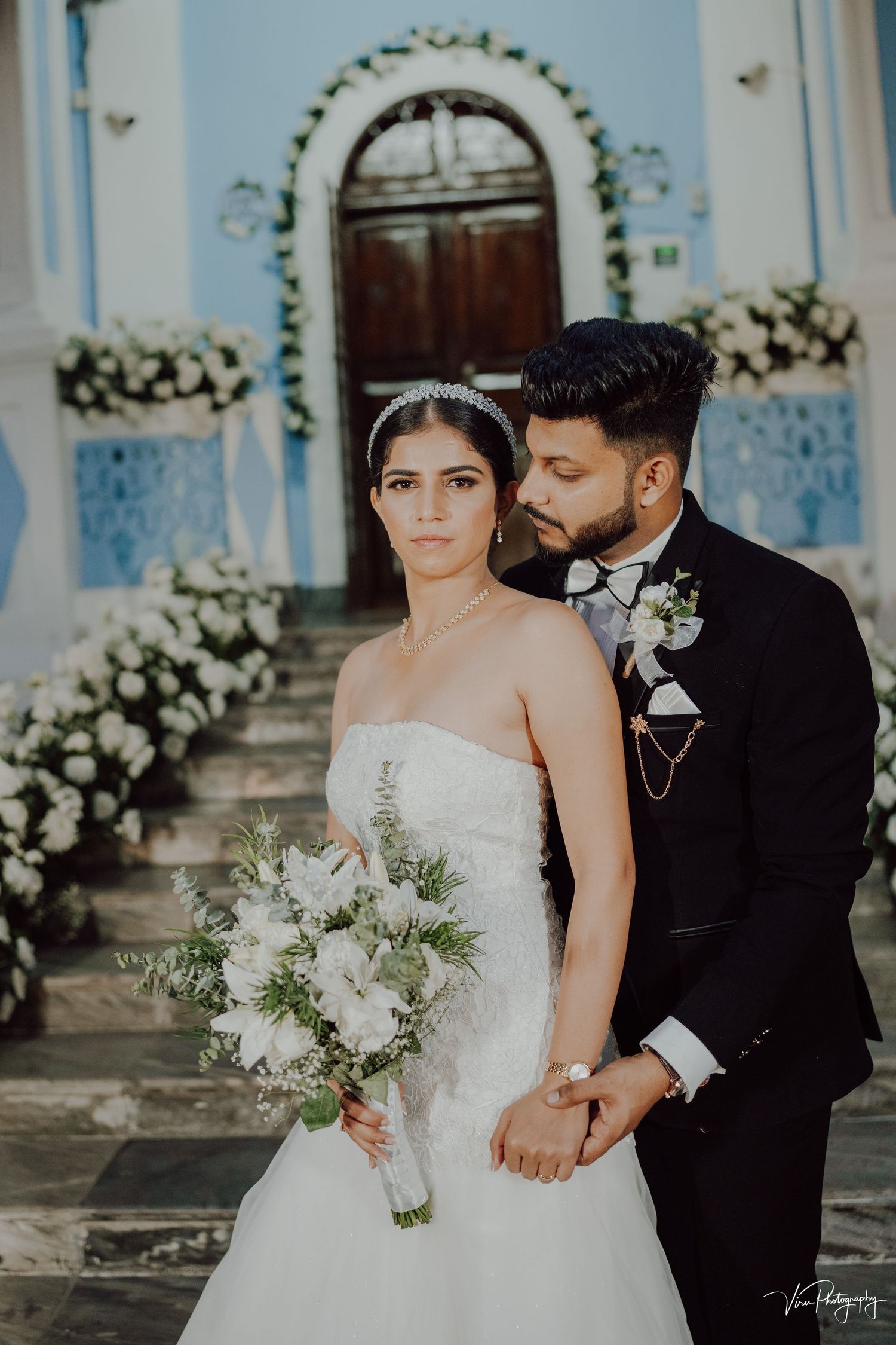 A couple in wedding attire poses on stone steps in front of a blue church with white floral decorations.