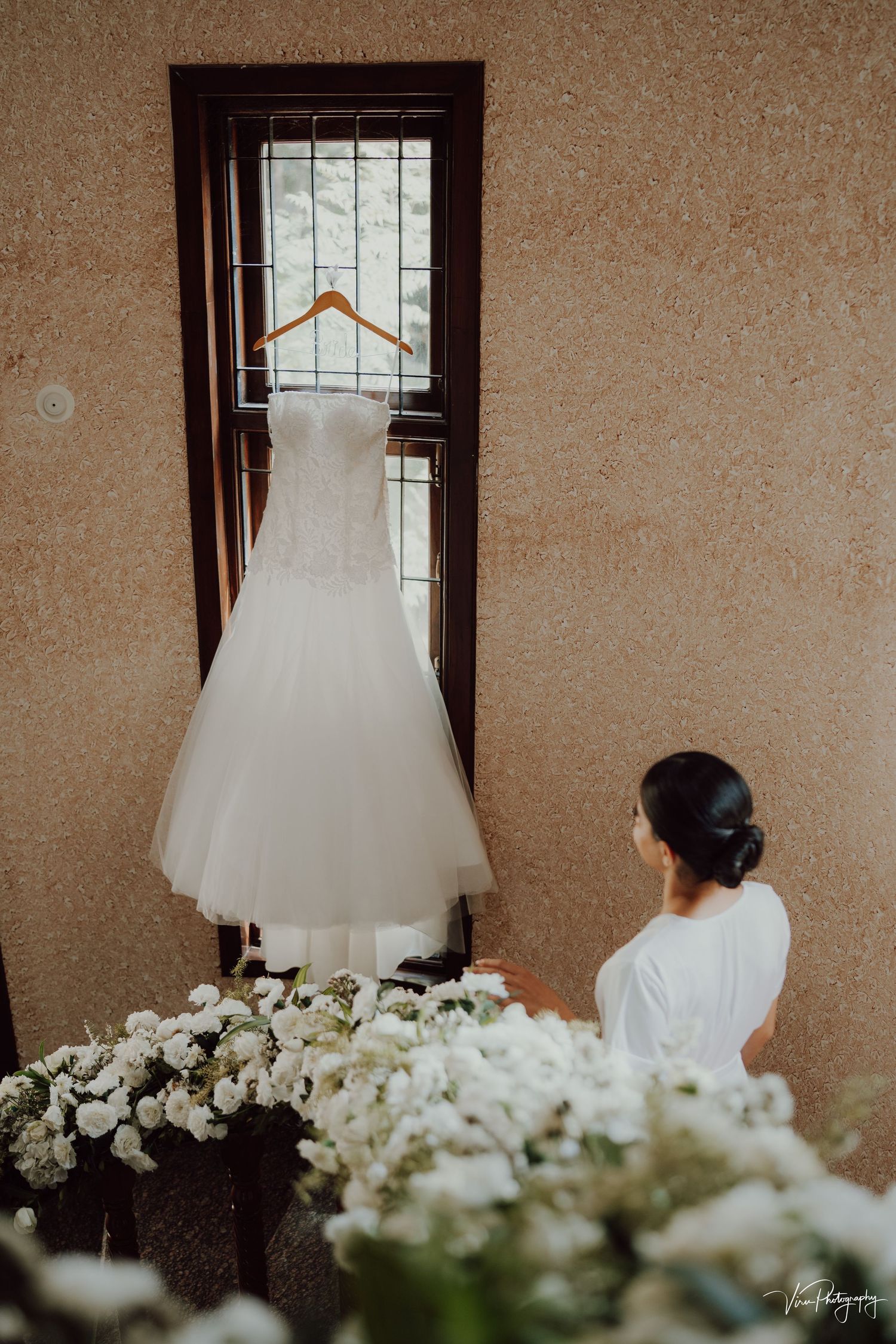 A white tulle wedding dress hangs in a window beside a floral arrangement in a moody, intimate setting.