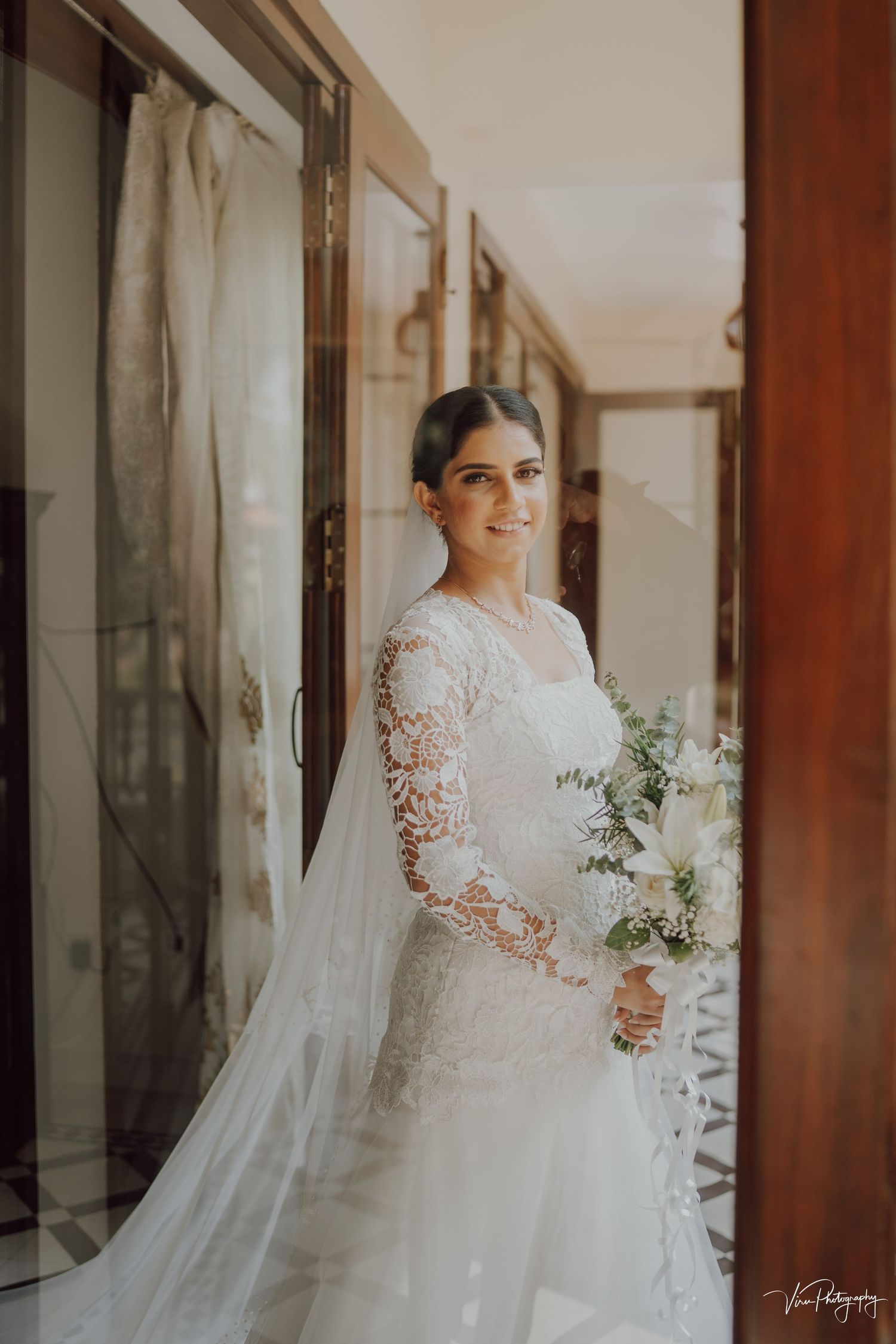 A bride in an elegant long-sleeved lace wedding gown holds a delicate bouquet of eucalyptus in a sunlit hallway.