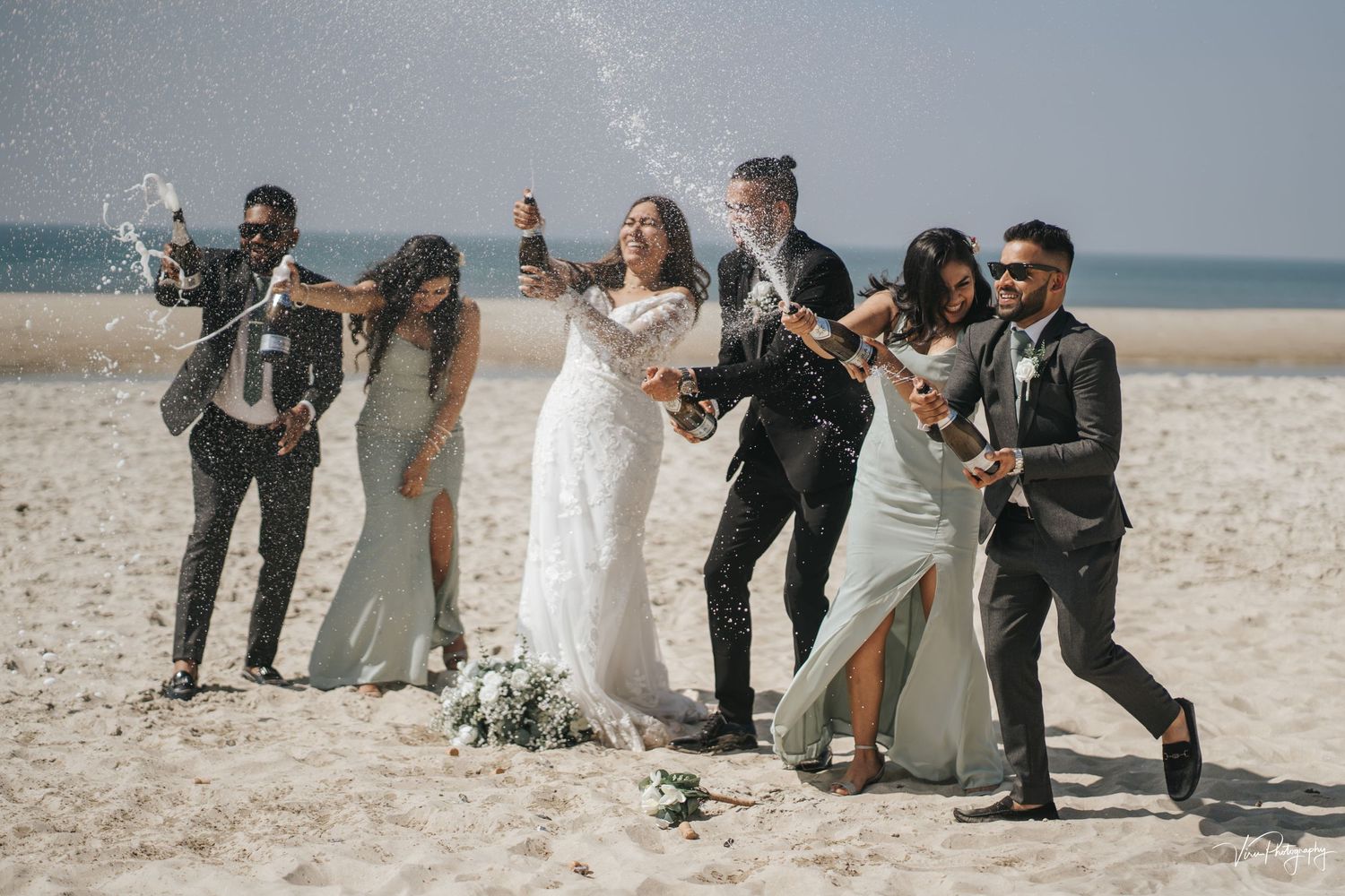 Wedding party celebrates joyfully on a sunny beach with champagne and flowers against a hazy ocean backdrop.