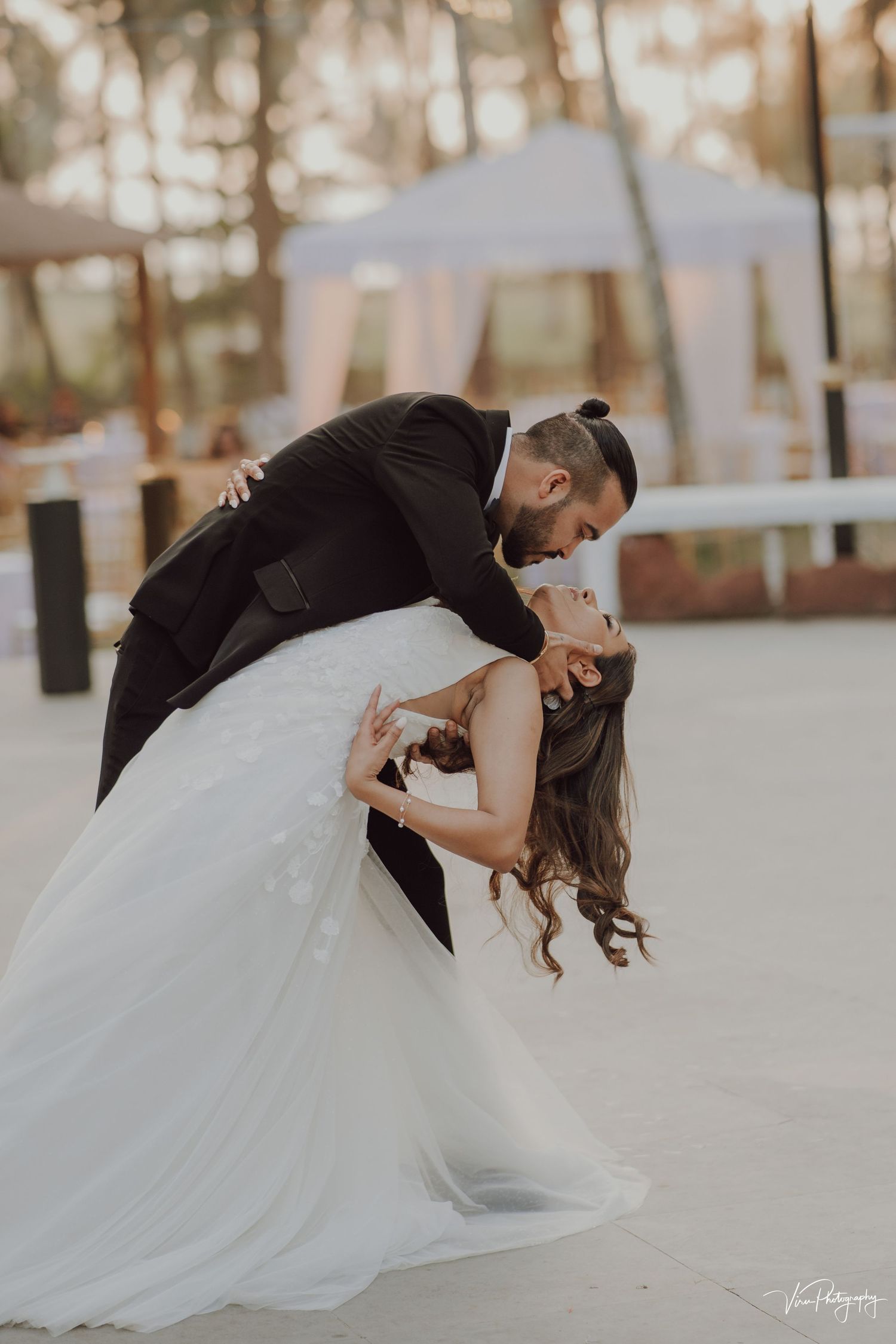 Romantic couple in a dip pose at an outdoor wedding venue with tents in the background at sunset.
