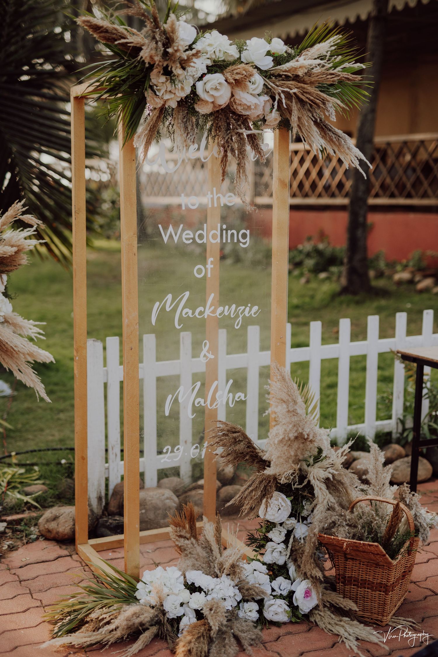 Elegant wedding welcome sign with gold frame, white flowers, pampas grass and greenery decorations on outdoor lawn setting.