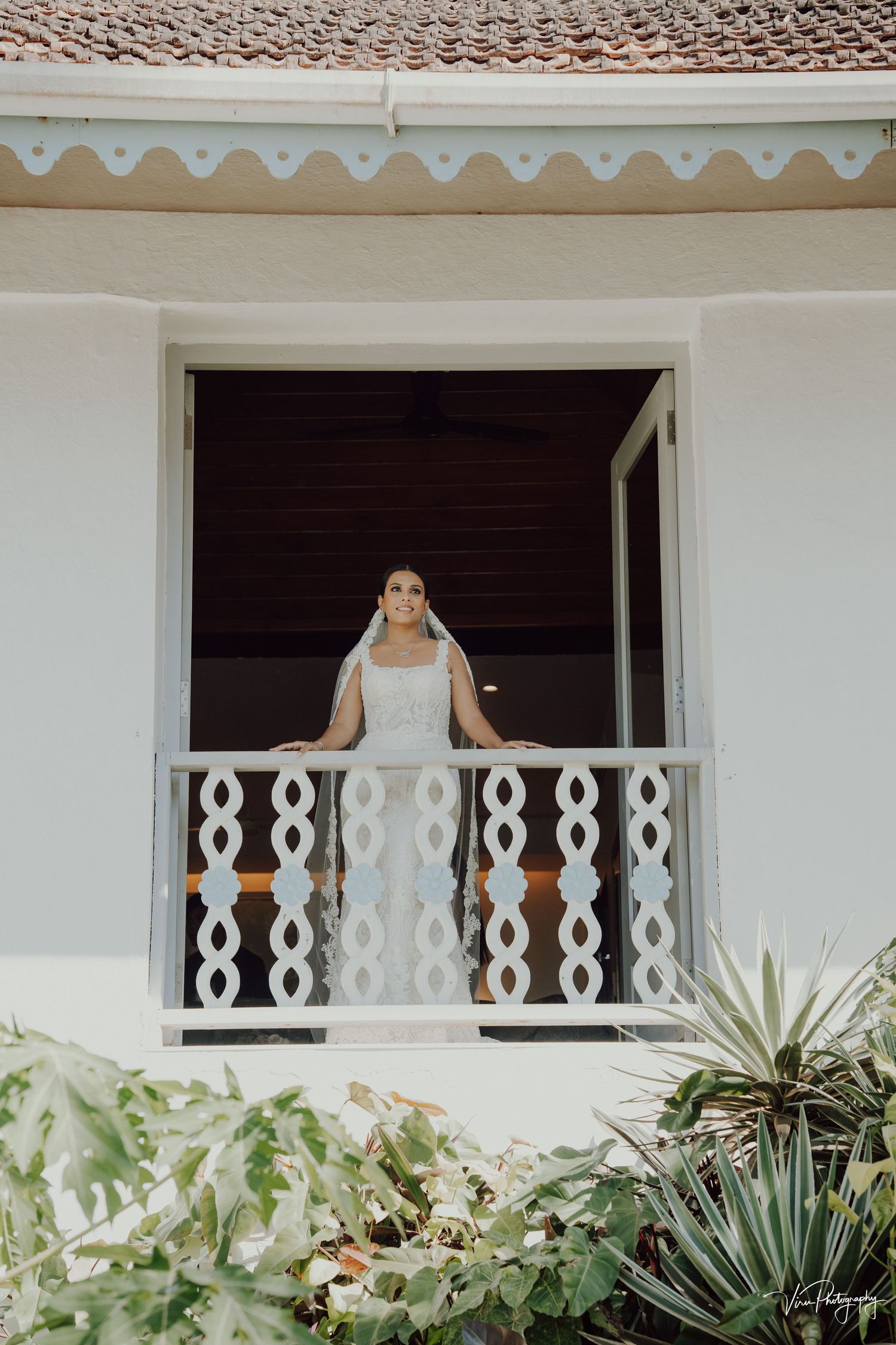 A white wedding dress is framed by an ornate balcony railing with tropical plants below at a Spanish-style venue.