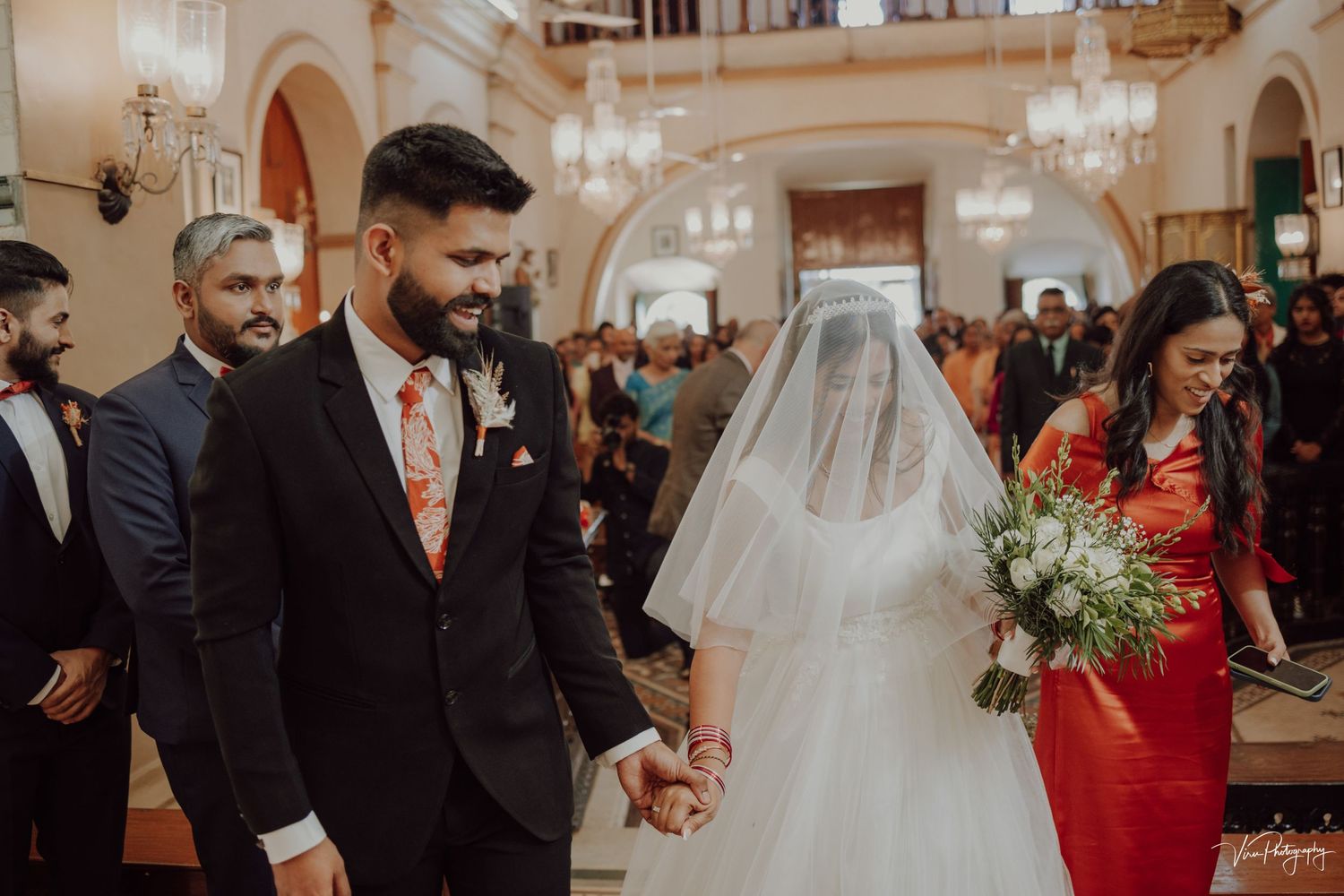 A couple walks down the aisle during their wedding ceremony in an ornate church with elegant chandeliers.