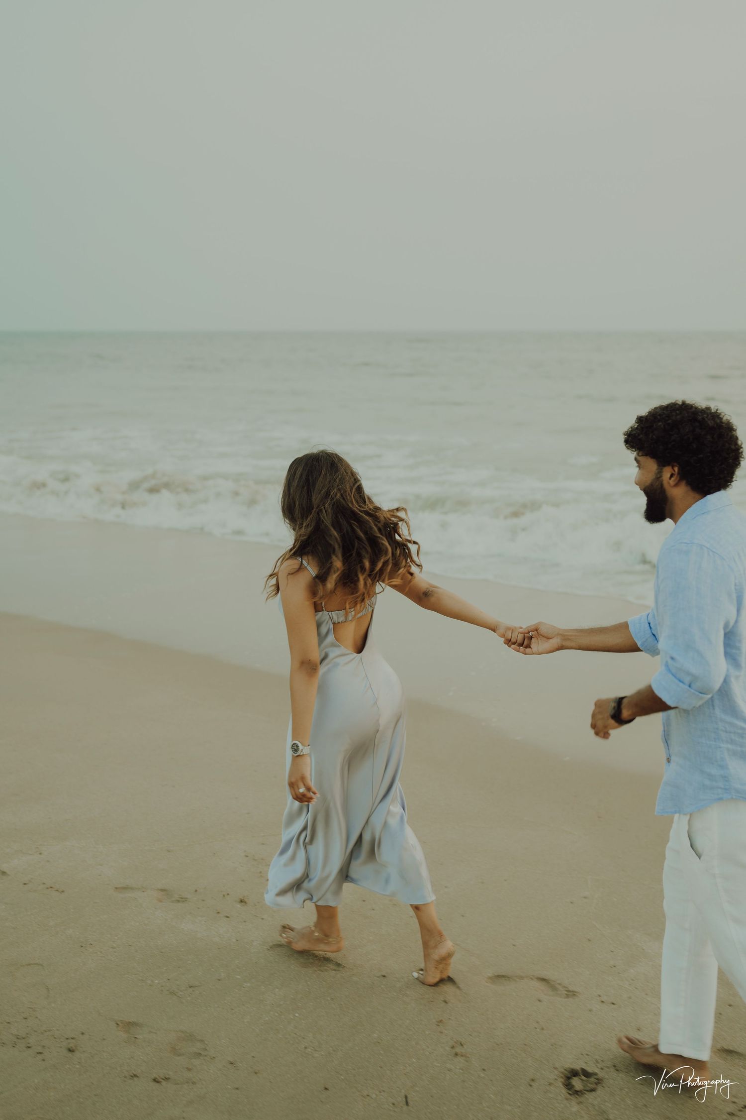 Couple holds hands while walking along misty beach at sunset in romantic ethereal scene.