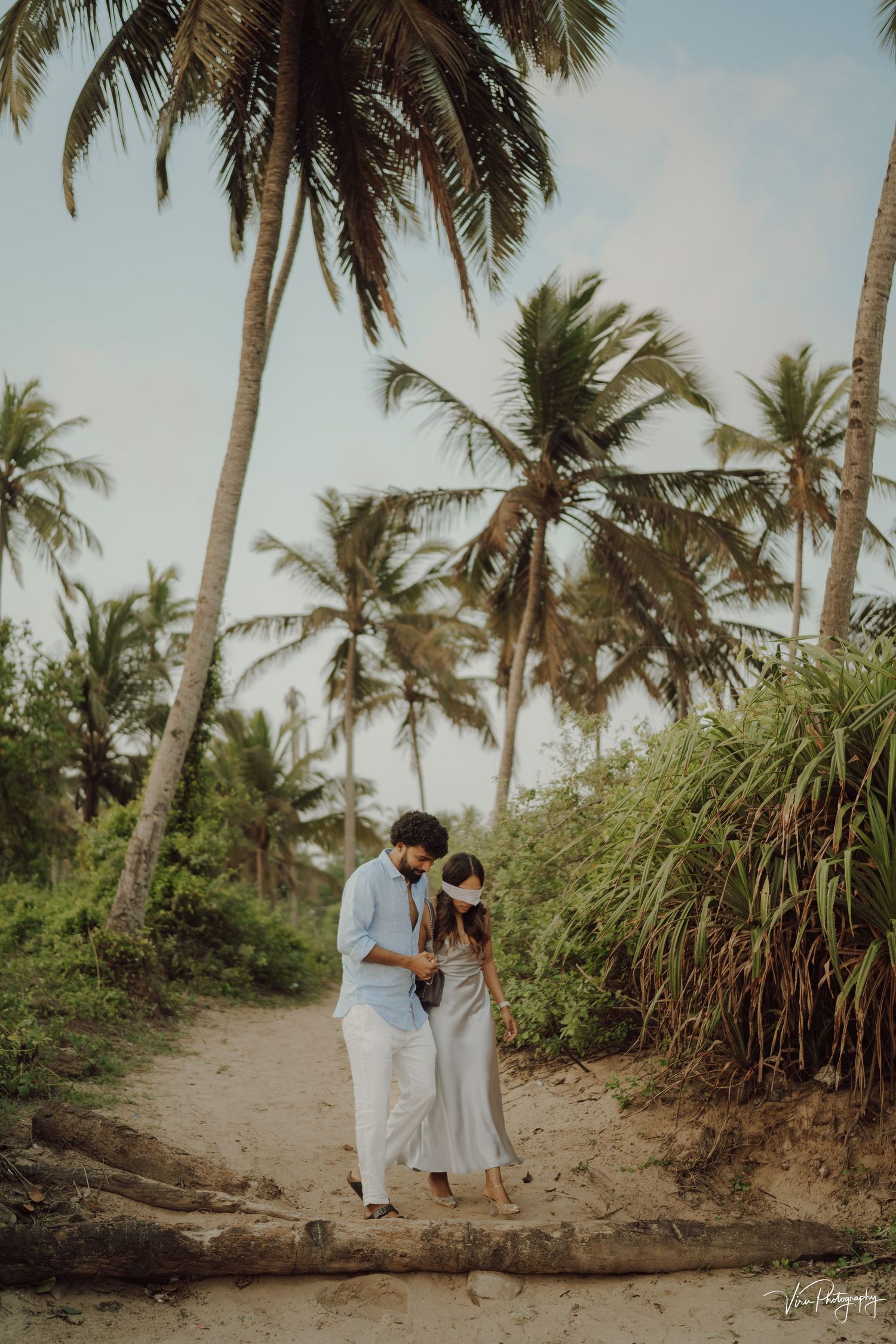 Romantic beach scene with couple embracing on sandy path surrounded by tropical palm trees in golden light.