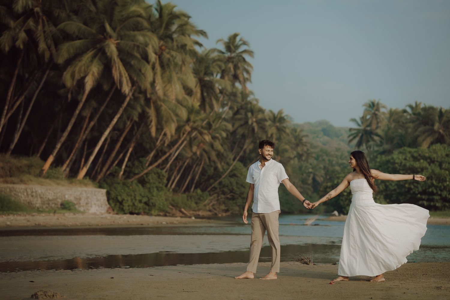 Romantic beach wedding photoshoot with palm trees and ocean waves along a tropical shoreline at sunset.