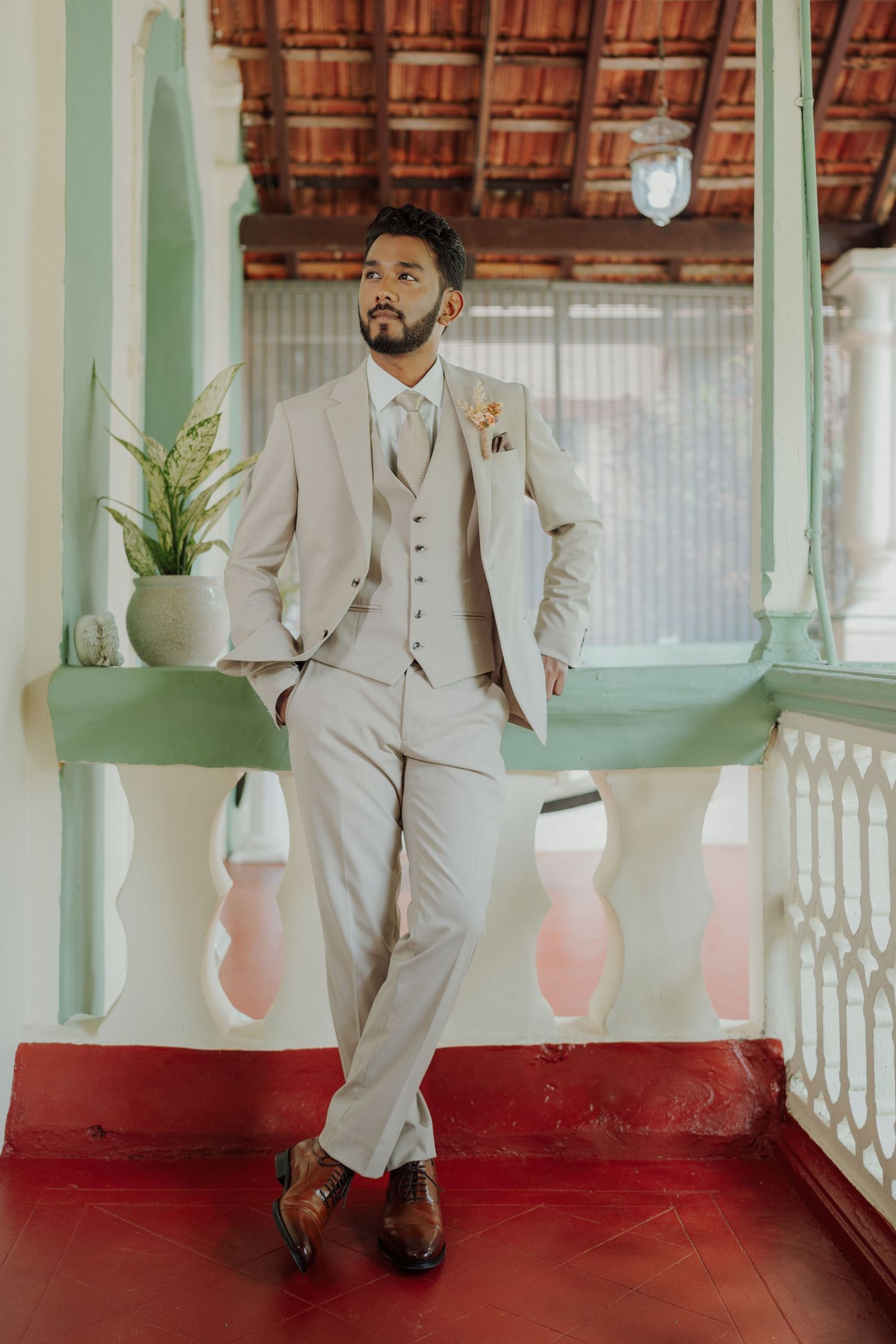 Groom in a beige three-piece suit and brown shoes leans on a green balcony railing with a red tiled floor.